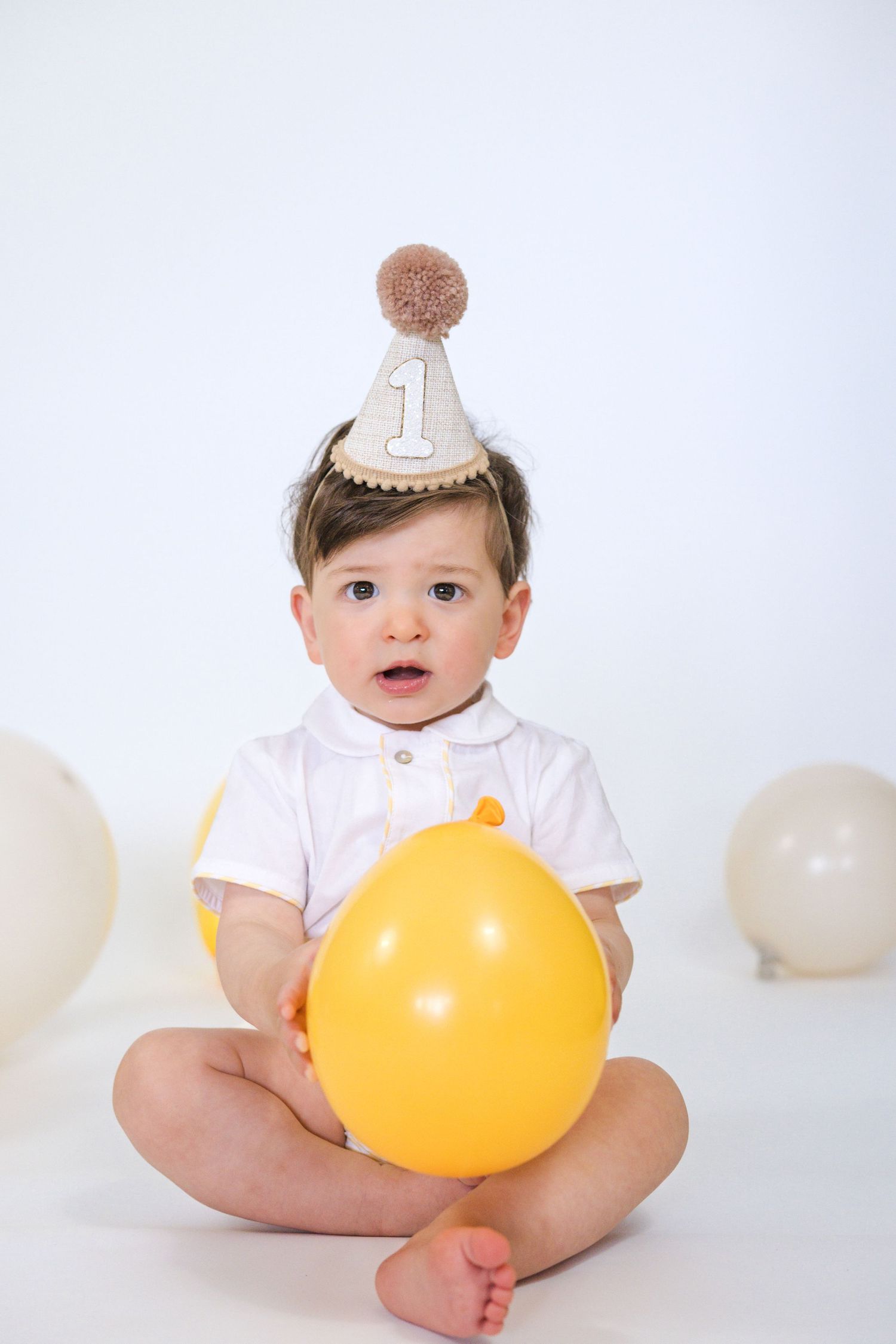 Baby wearing party hat holds yellow balloon against white backdrop.