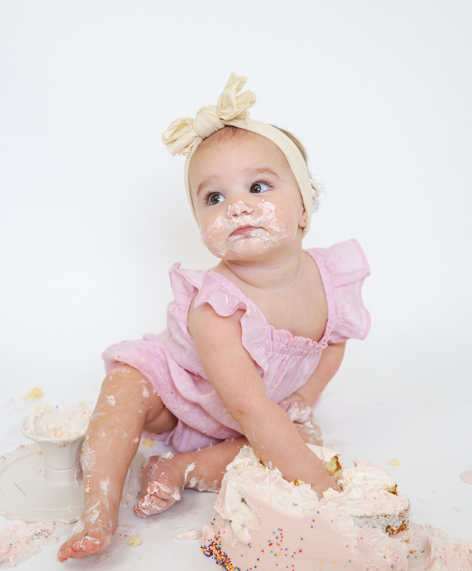 Baby in pink dress crawling through destroyed cake during first birthday photoshoot.