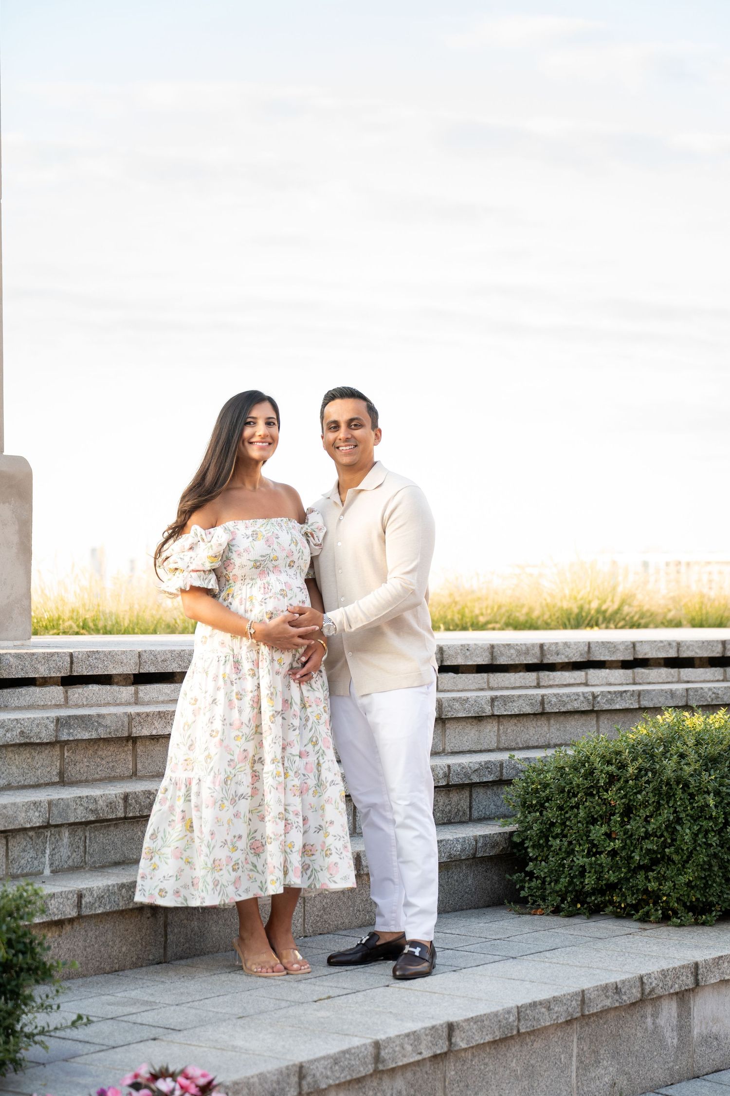 A romantic photoshoot on stone steps with a couple in white and floral attire against a bright sky.
