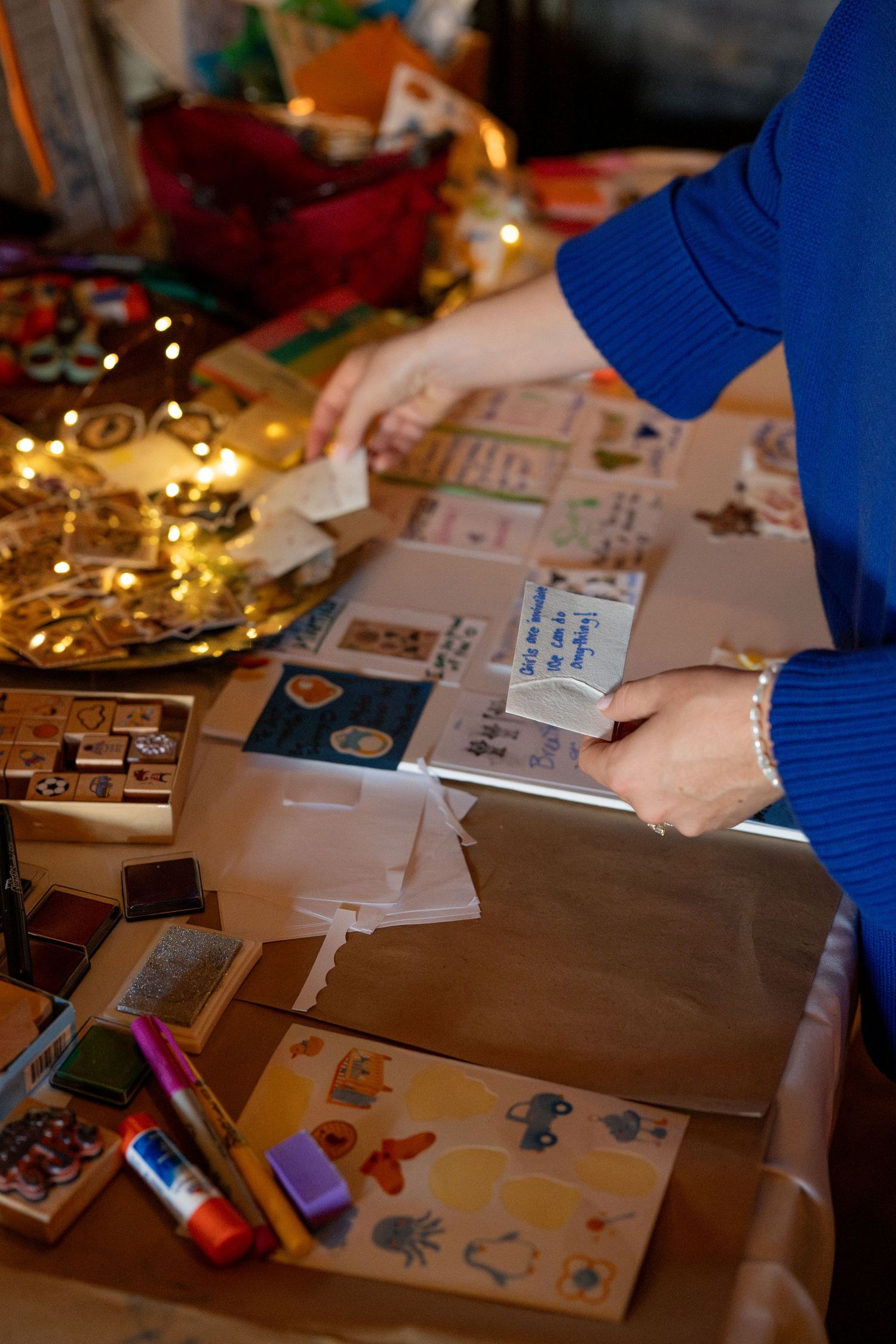 Hands work on wrapping presents and crafts at a table decorated with holiday lights and festive materials.
