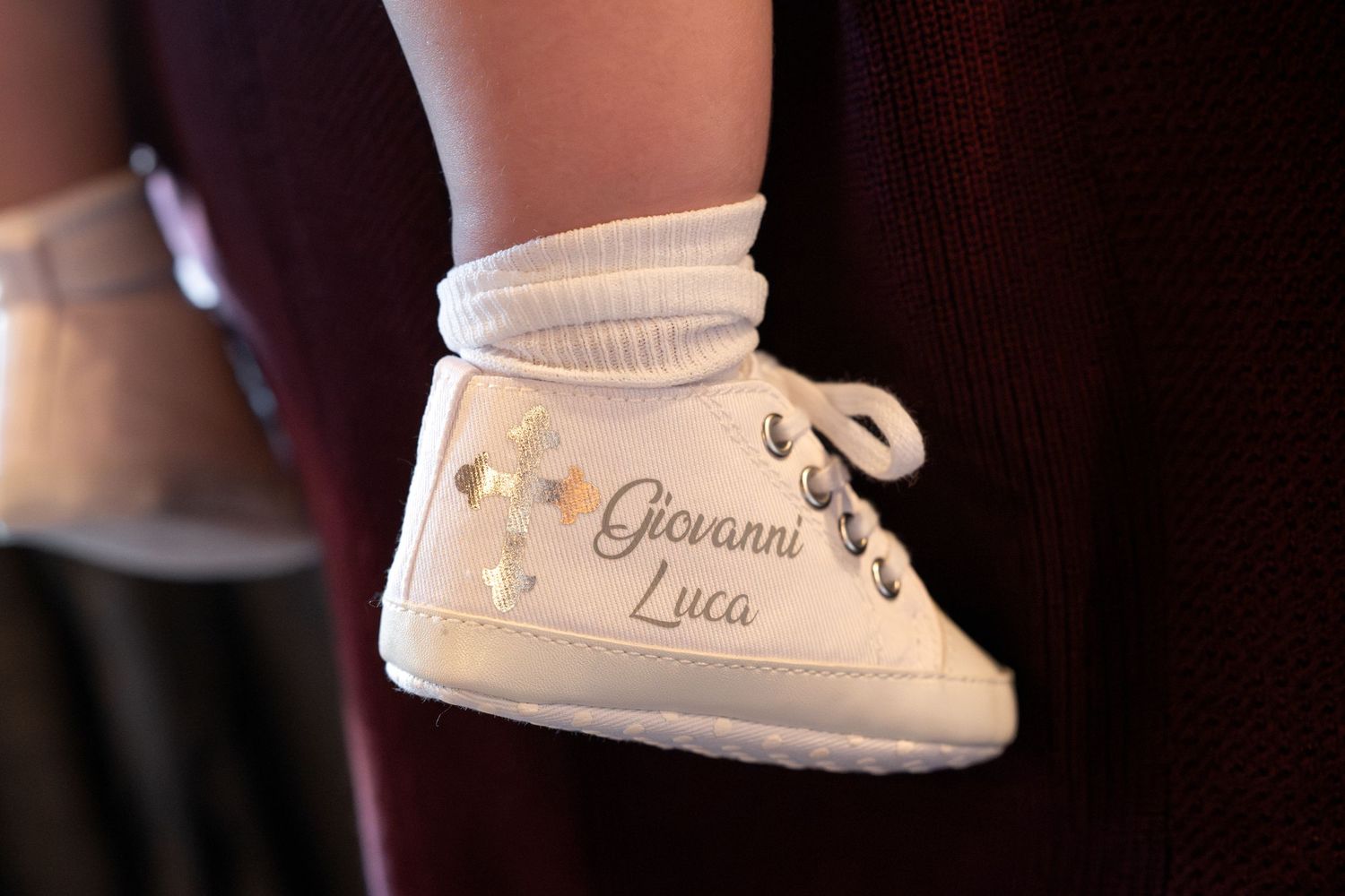A small white baby shoe with embroidered details and a white sock, photographed in soft lighting.
