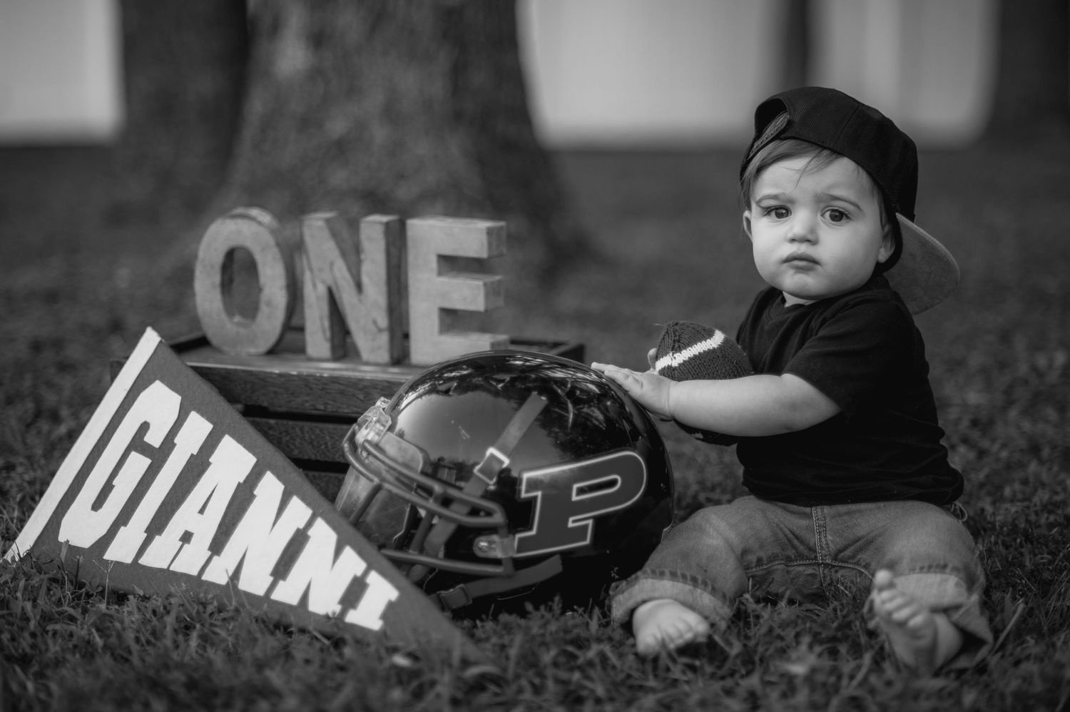 Baby wearing a black baseball cap lying on grass with sports equipment.
