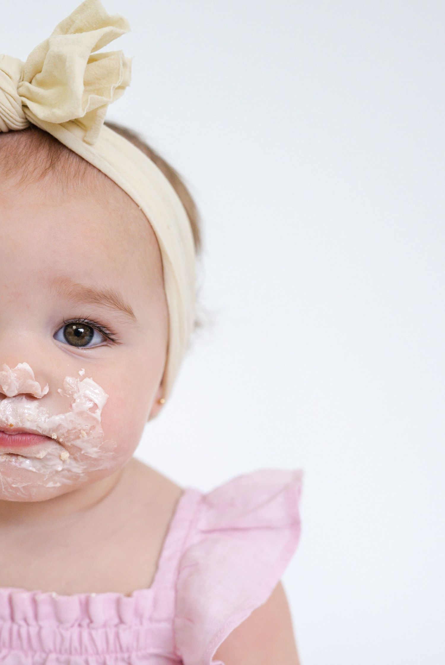 Close up of baby in pink dress and cream bow with cake frosting on face.