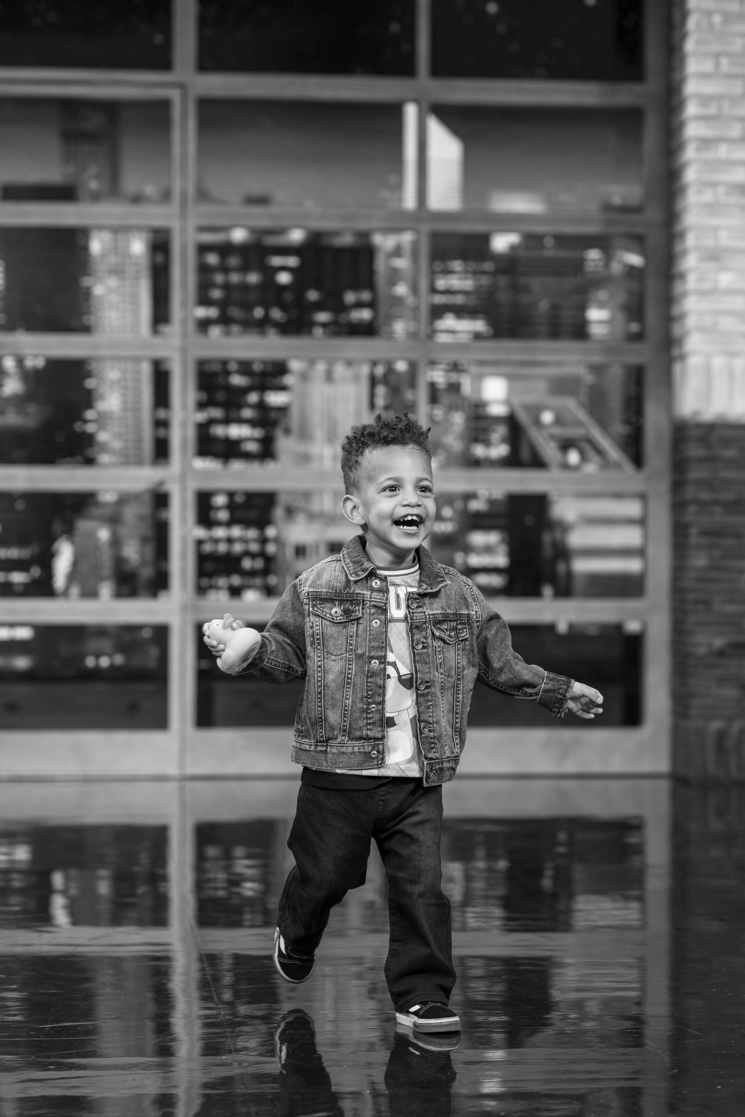 A joyful young boy in a denim jacket laughs and runs outdoors in a black and white urban photo.