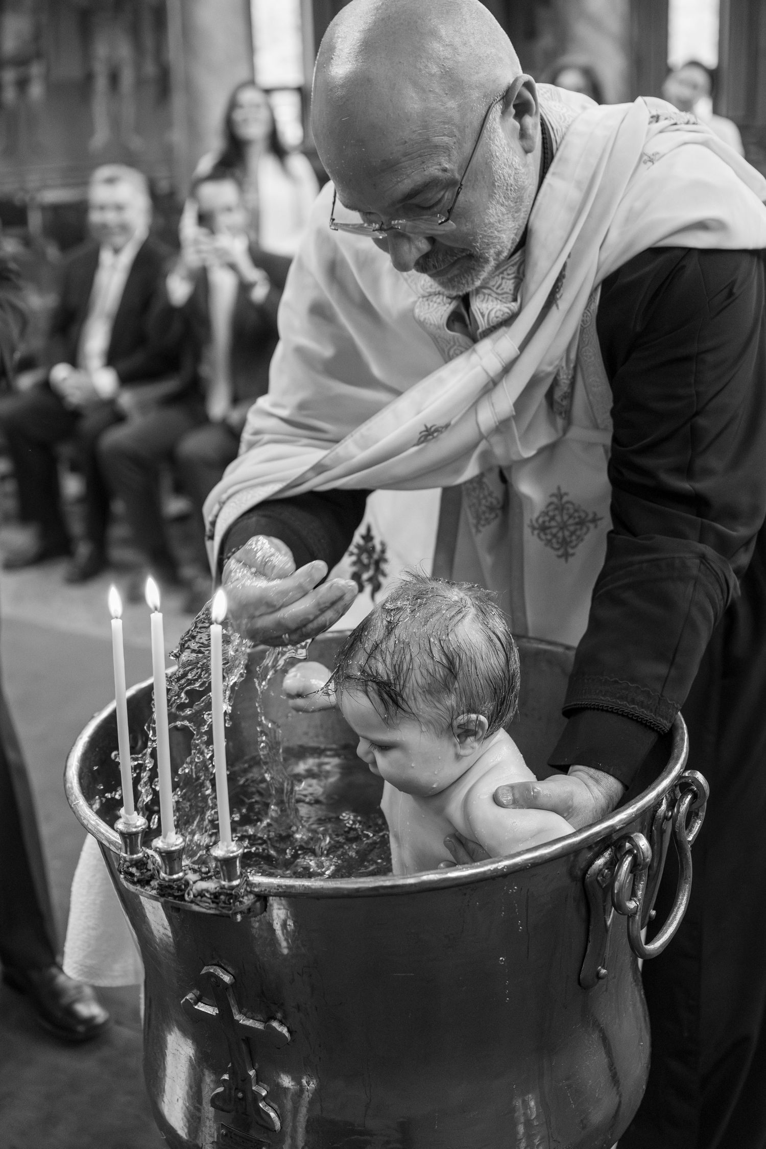 Priest performing Orthodox baptism, immersing infant in large font with candles, black and white church ceremony photo.