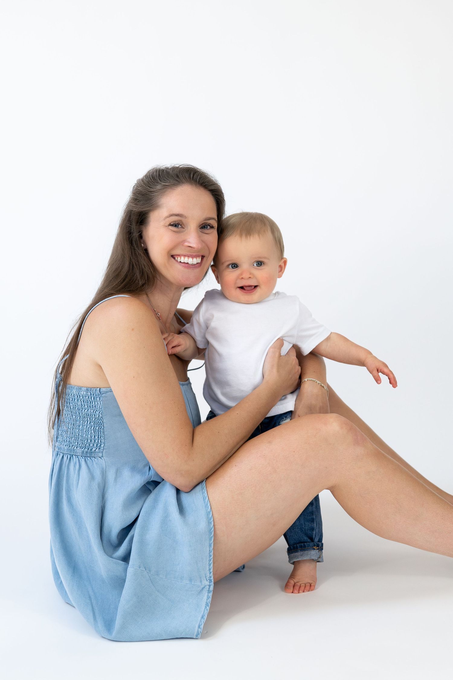 Smiling mother in blue dress sitting on white background holding happy baby in white onesie and jeans.