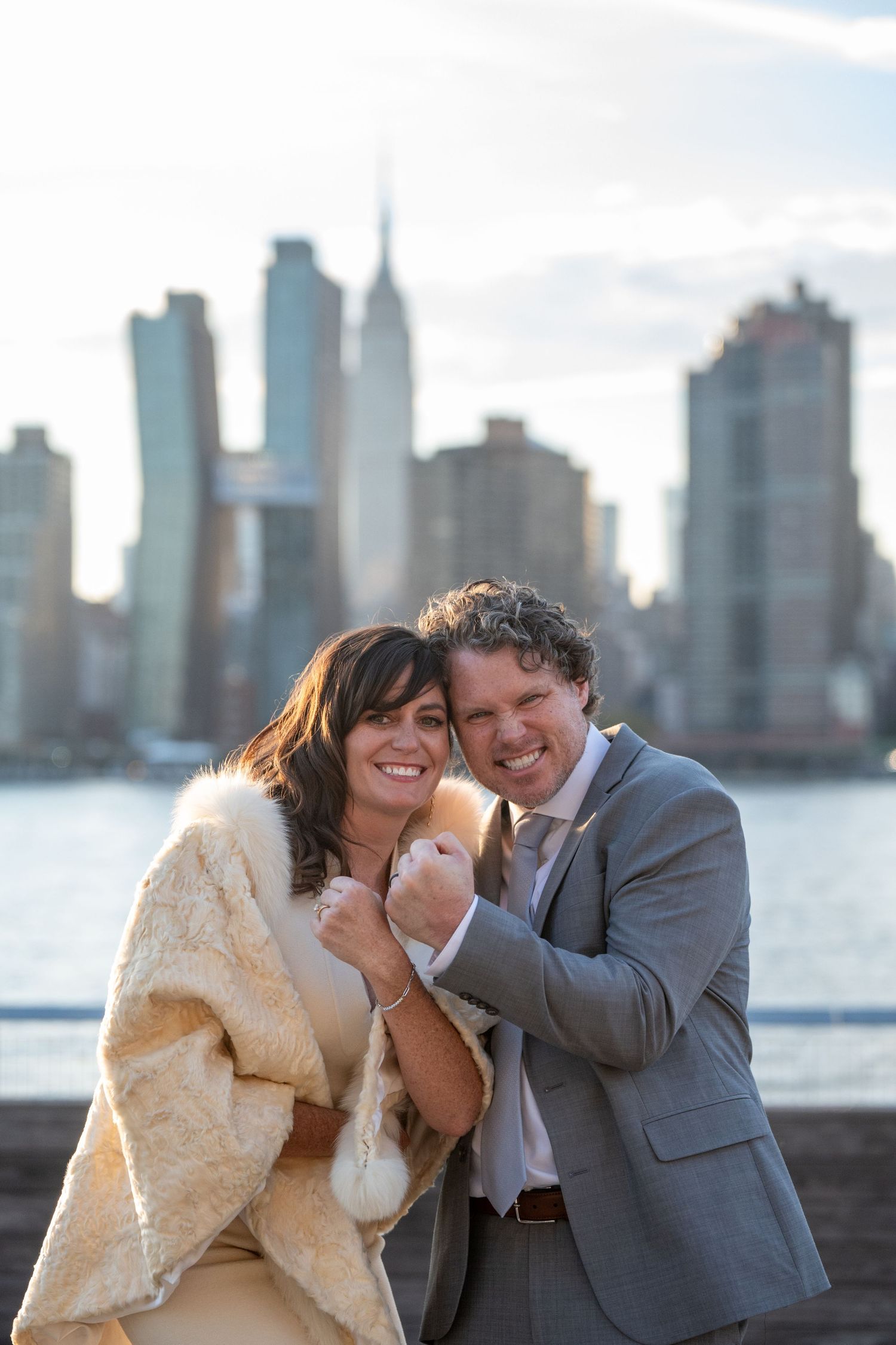 A romantic couple shares a joyful moment along the waterfront with the New York City skyline in the background.