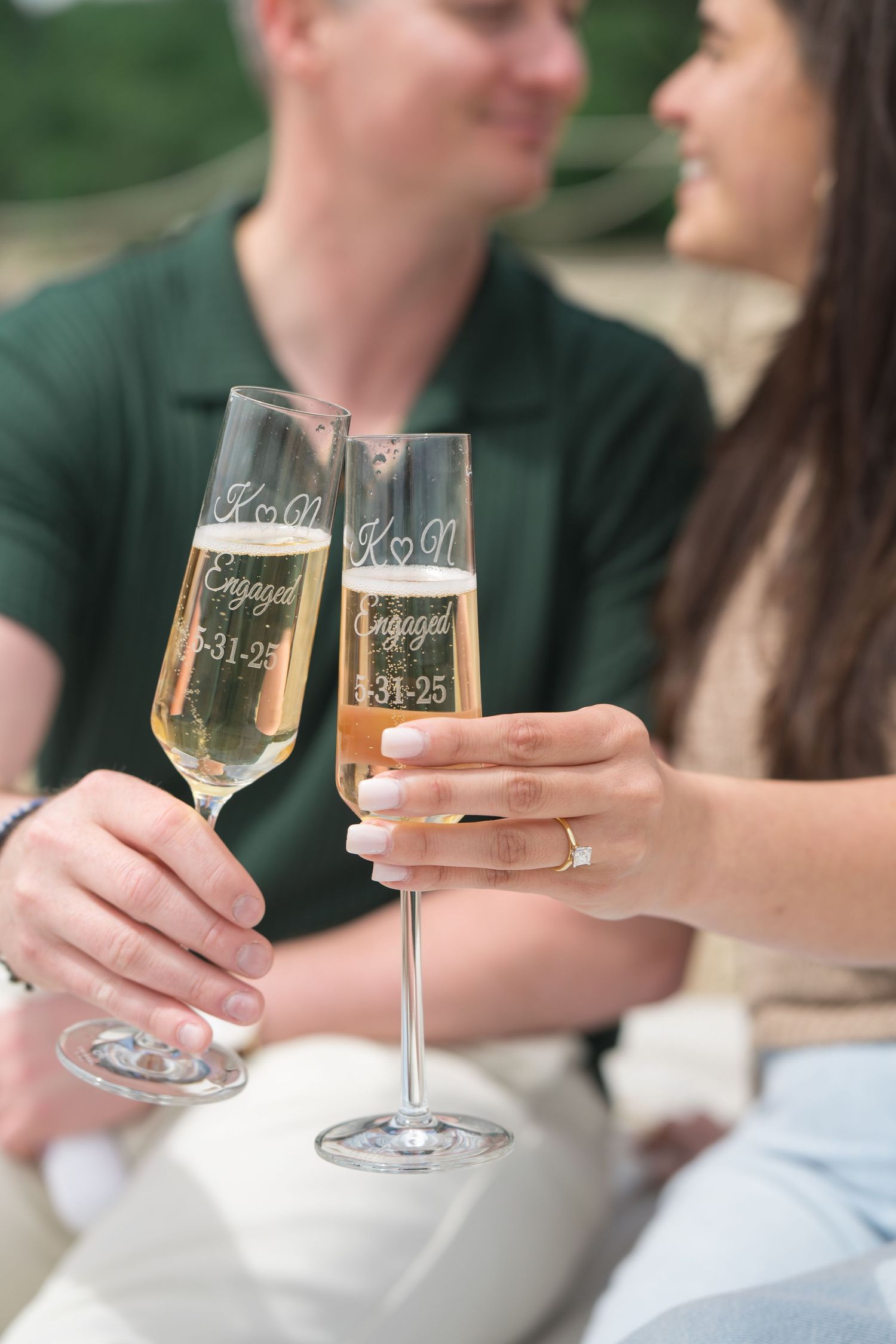 Two champagne flutes clink together in celebration with an engagement ring visible.