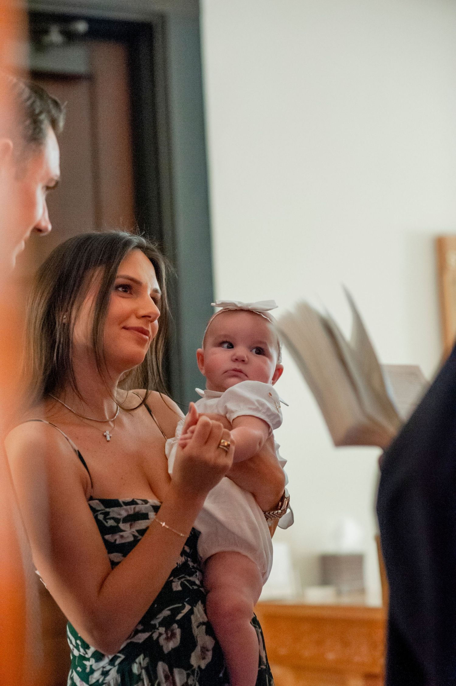 Woman in black floral dress holds and interacts with a smiling baby in a warm, sunlit room.