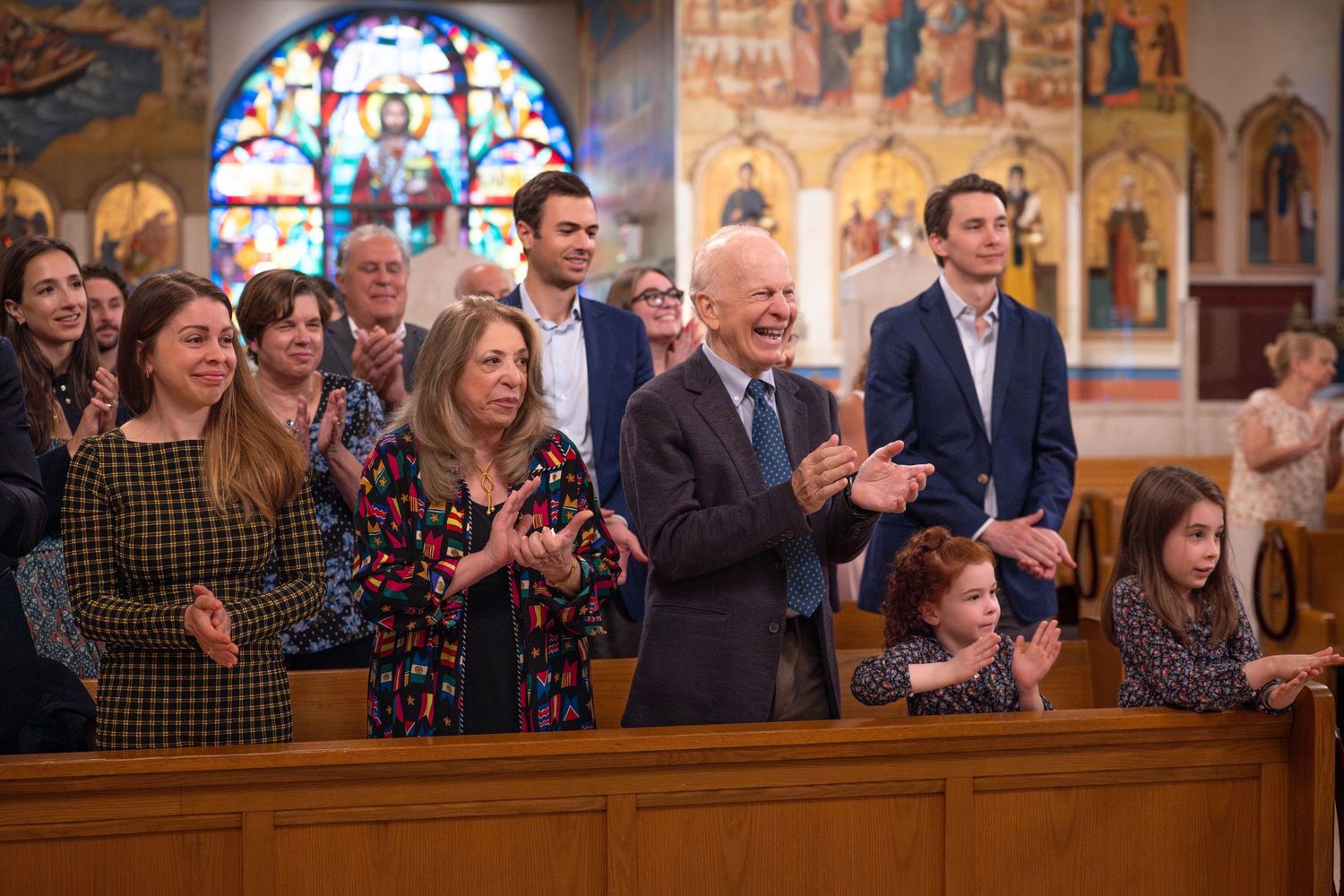 People gathered in church pews during a service with stained glass windows and religious artwork visible in background.