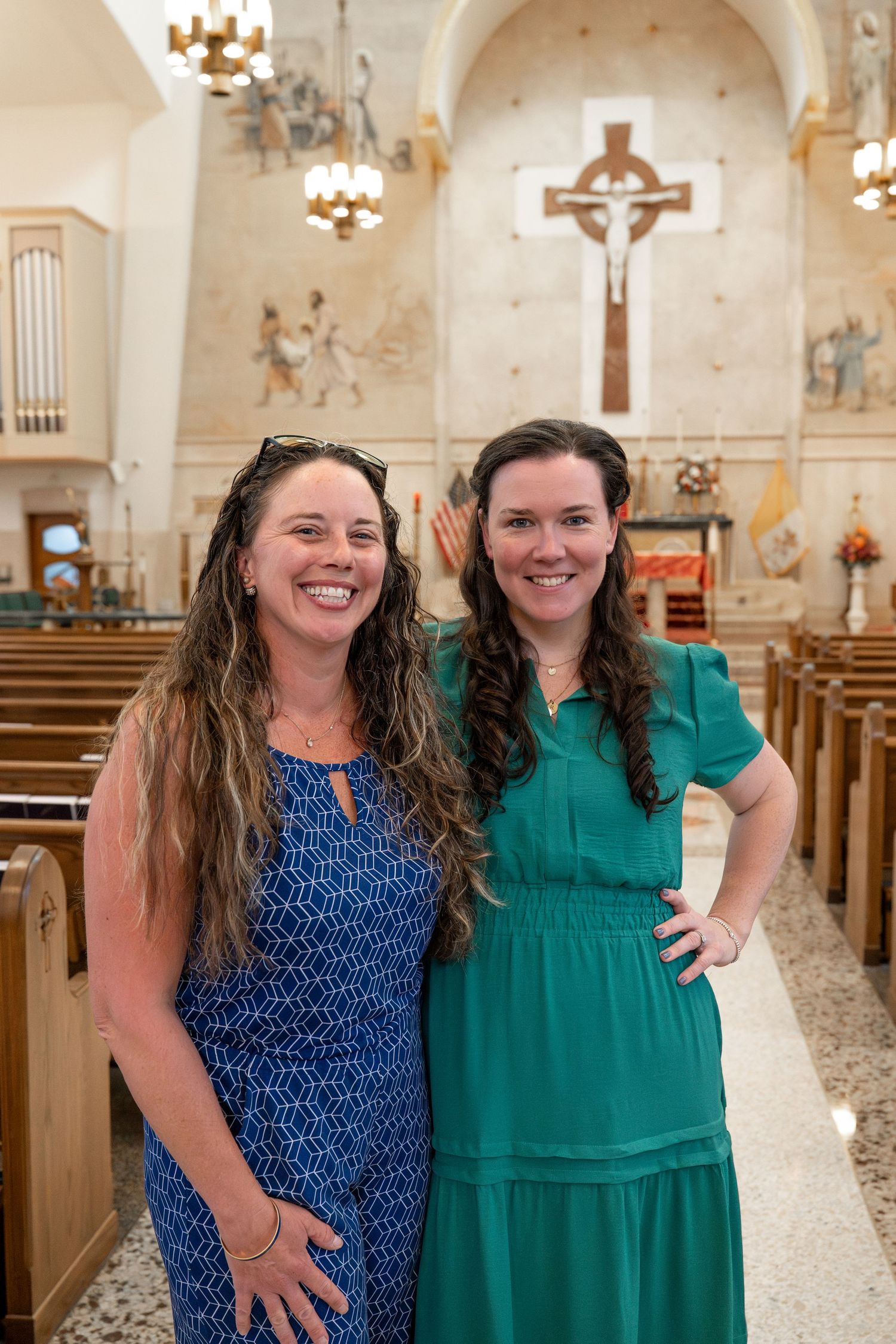 Two women wearing blue and green dresses smile together inside a church with a crucifix visible in the background.