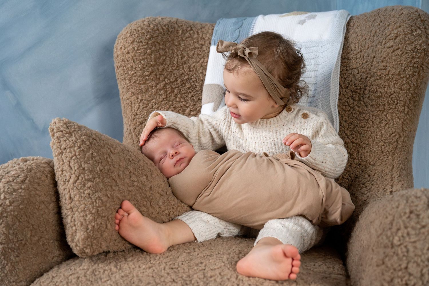 A sleeping baby wrapped in a white blanket rests peacefully on a brown plush couch.