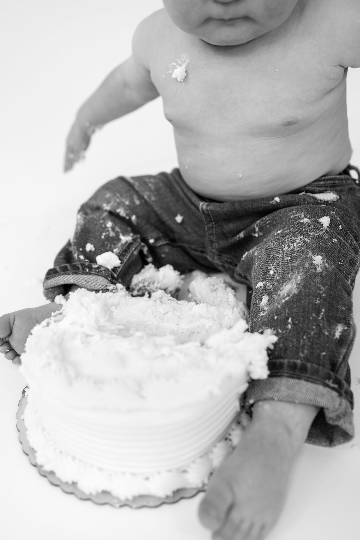 Baby in jeans smashing a white frosted cake, covered in icing, black and white photo.