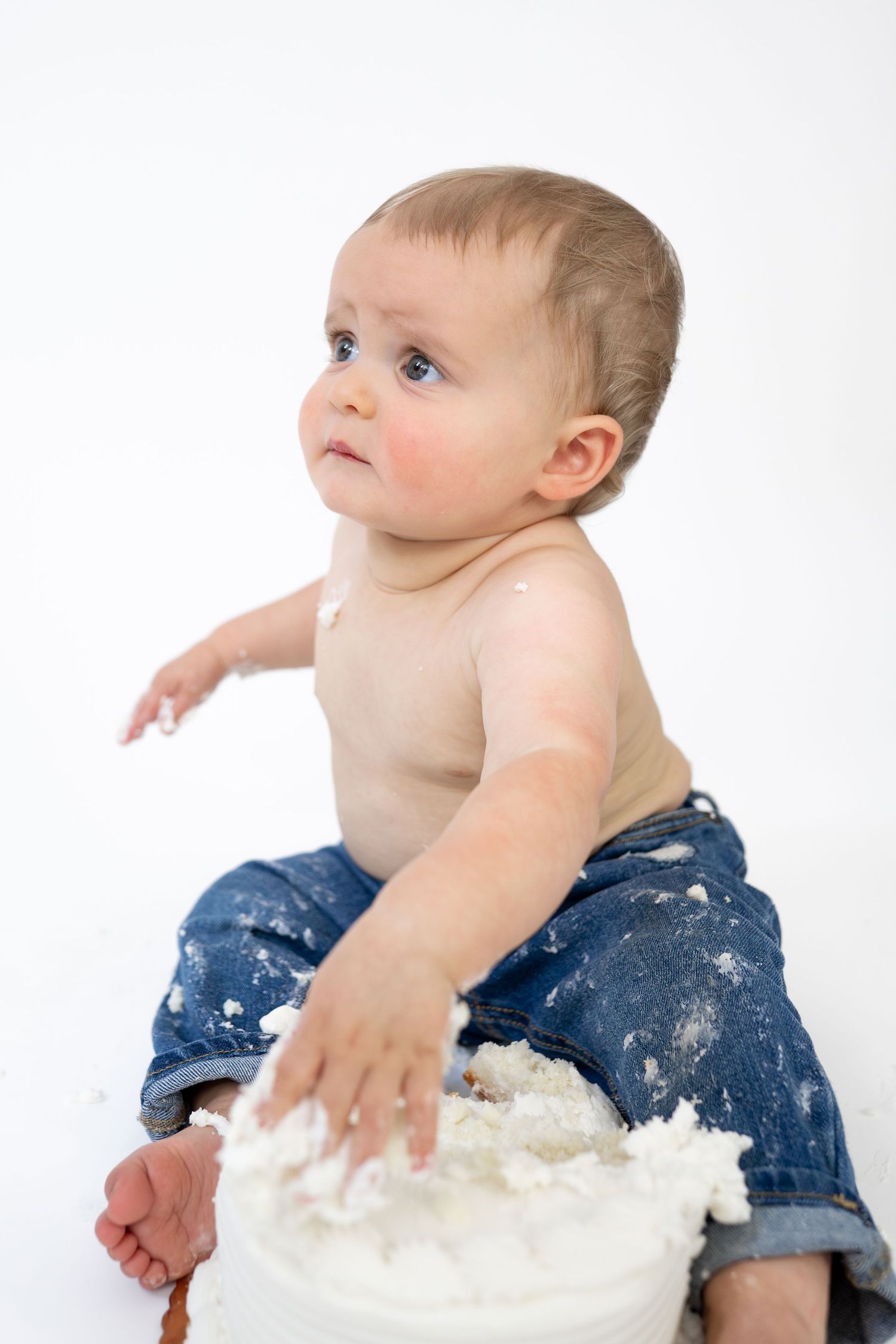 Baby in jeans smashing a white cake with frosting covered hands during a first birthday cake smash photo session.
