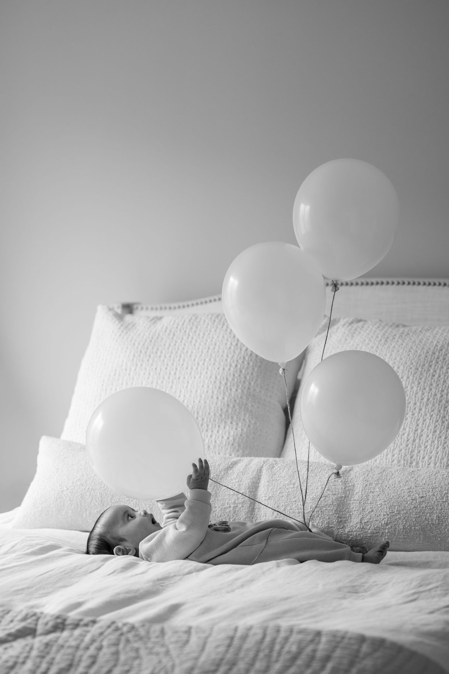 Black and white photo of a baby lying on a bed reaching for white balloons tied to the headboard.