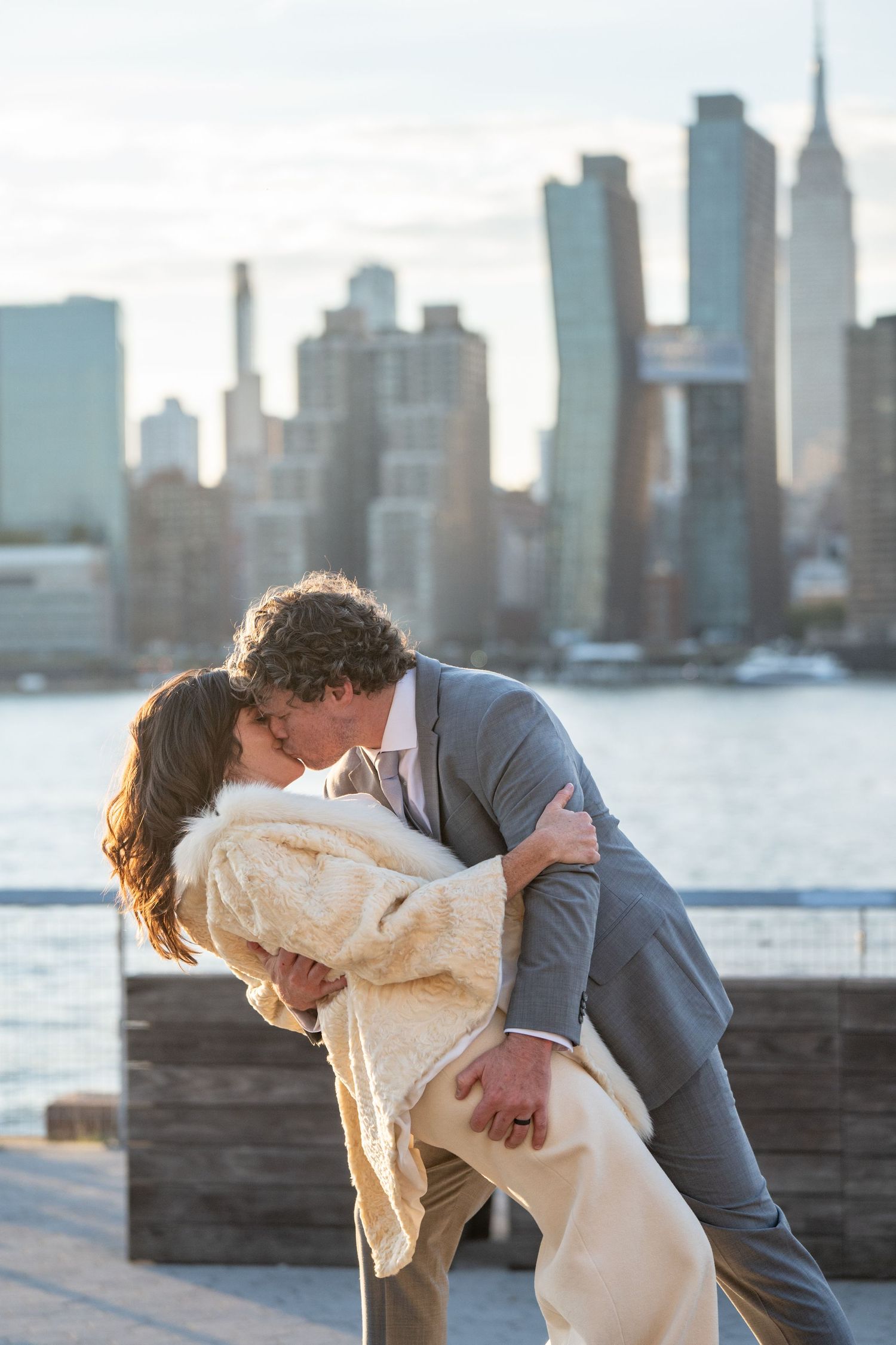 A romantic couple shares an intimate dip pose against the New York City skyline at sunset along the waterfront.