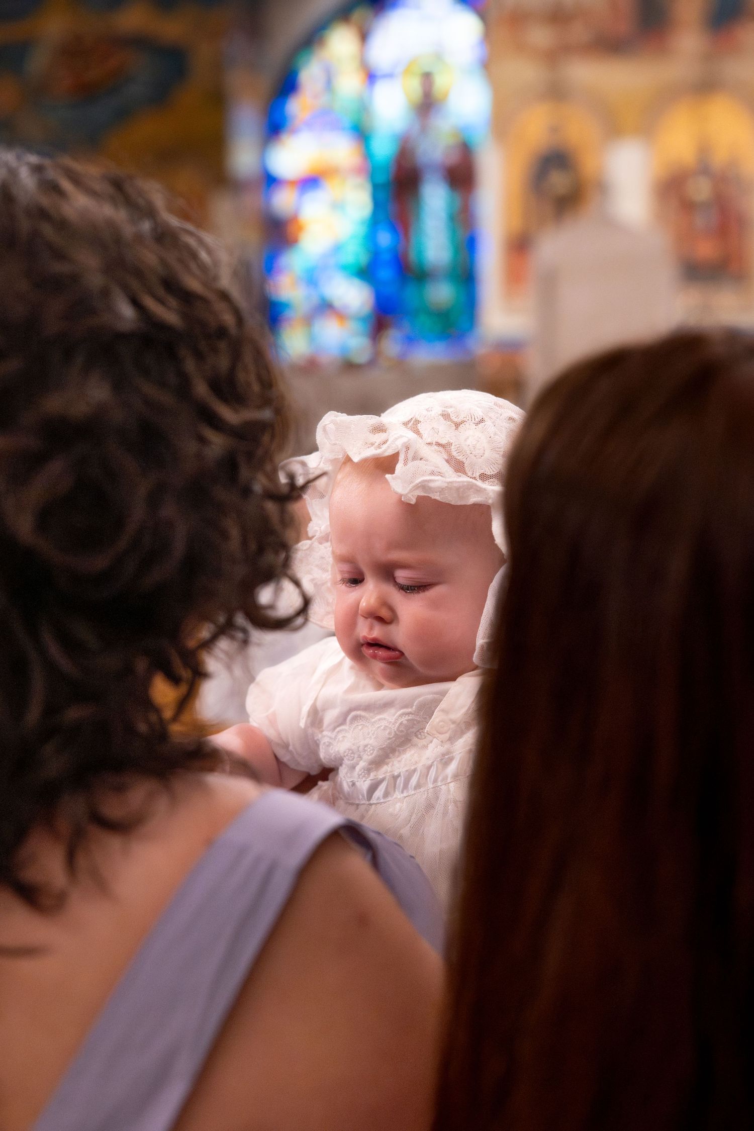 A baby wearing white baptismal clothing cries during the ceremony with stained glass windows in background.