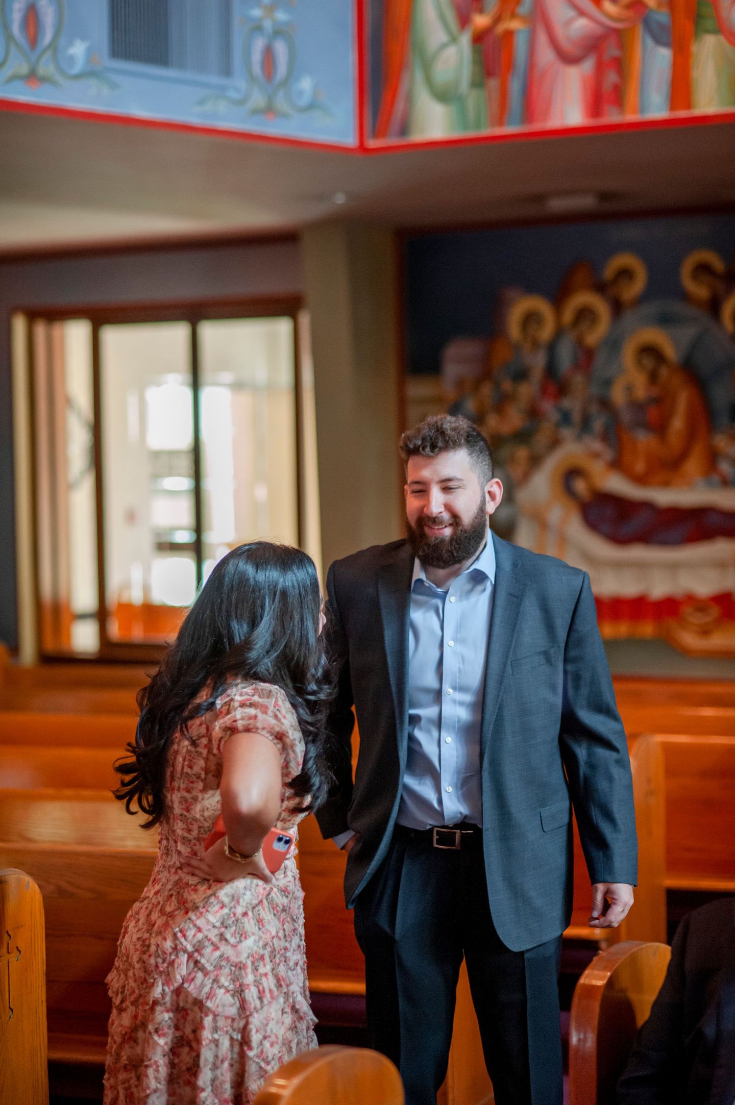 A couple shares a tender moment in a church with religious murals and wooden pews in the background.