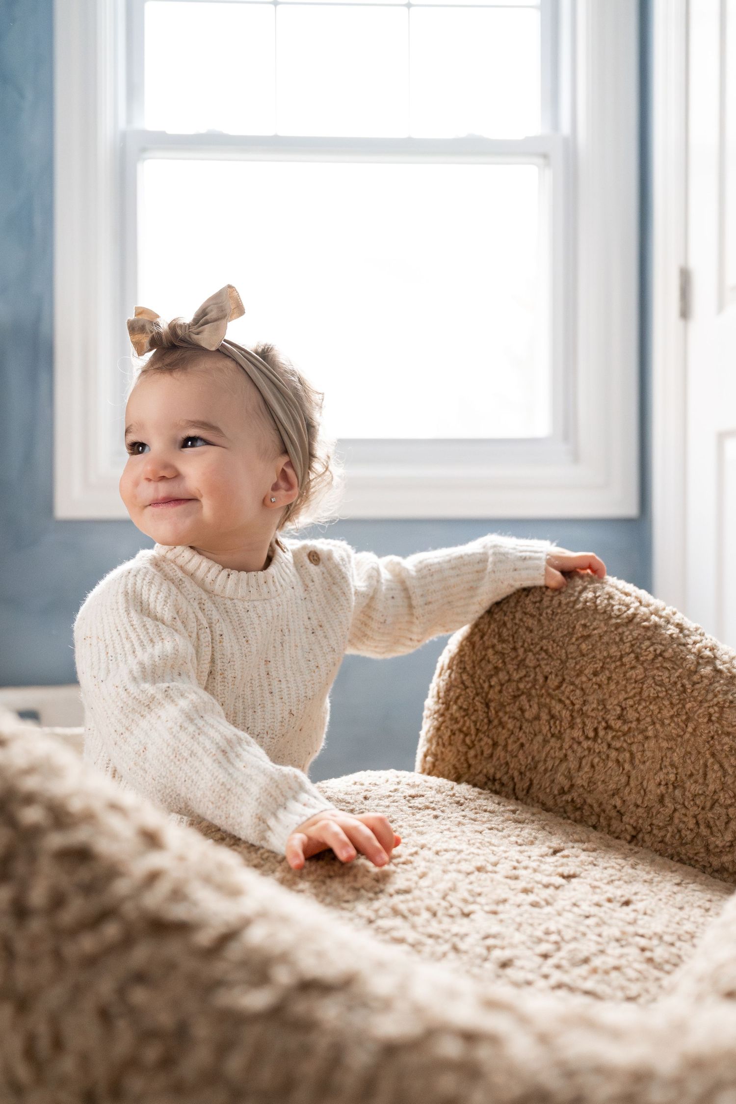 A small figure in a white sweater peeks over a plush beige couch in a bright room.