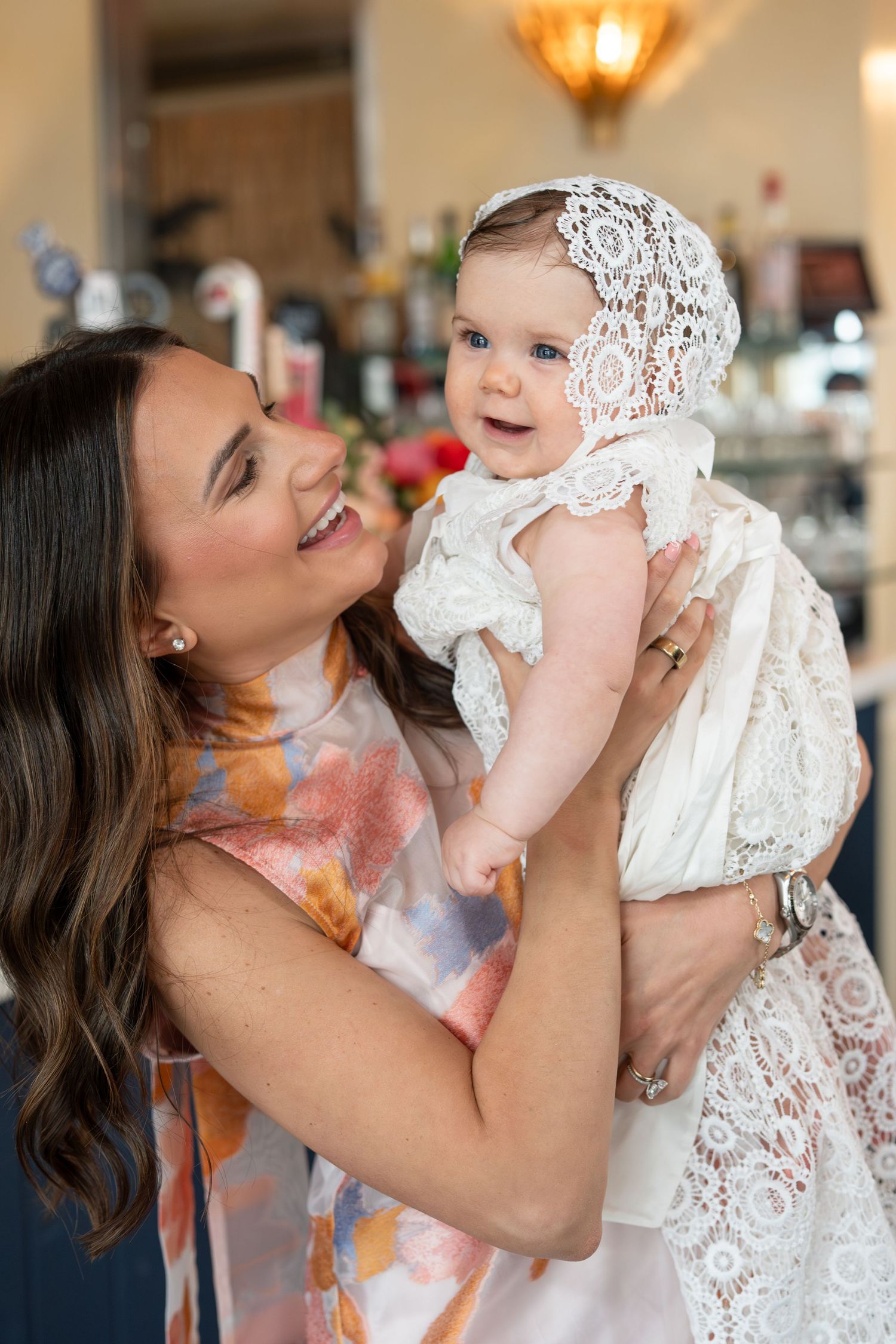 Joyful mother lifting smiling baby in white lace christening gown and bonnet.