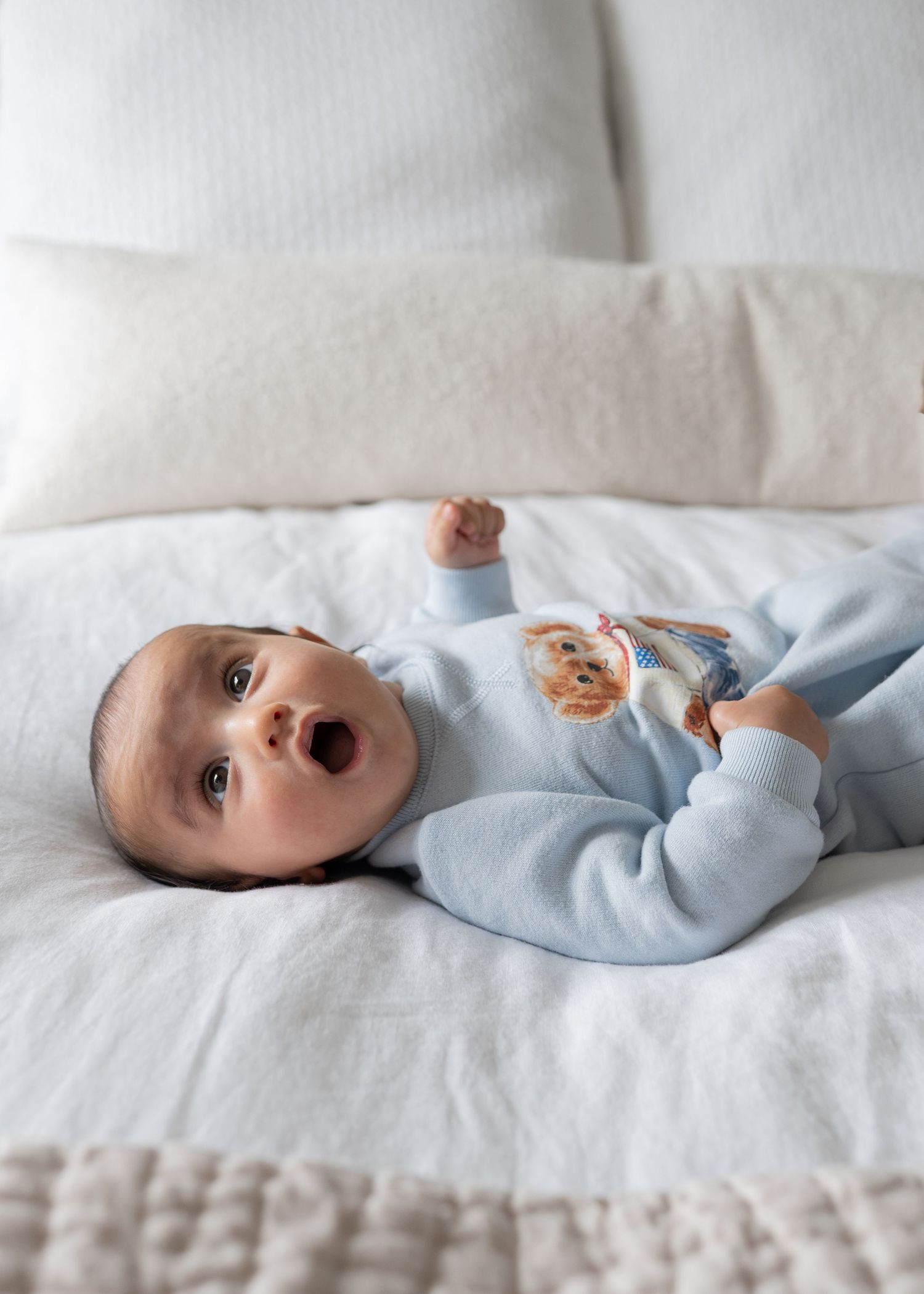 Baby in light blue bear romper lying on white bedding, mouth open.