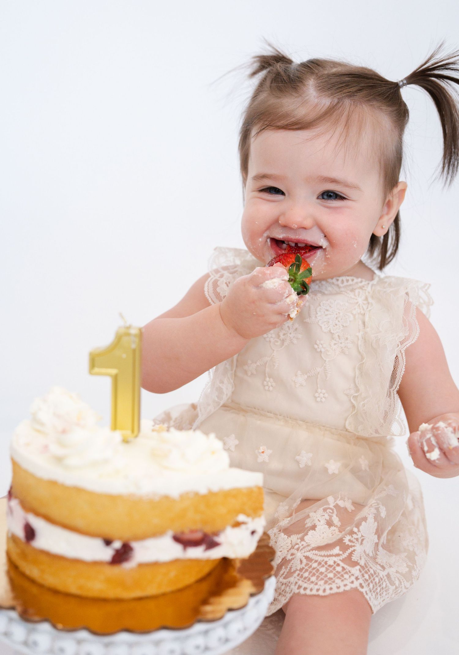 Baby in lace dress celebrating first birthday with cake and gold number one decoration.
