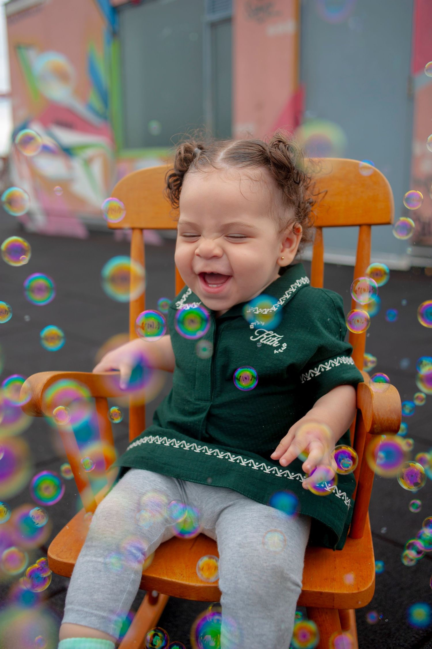 Child laughing in green dress while surrounded by soap bubbles in orange chair.