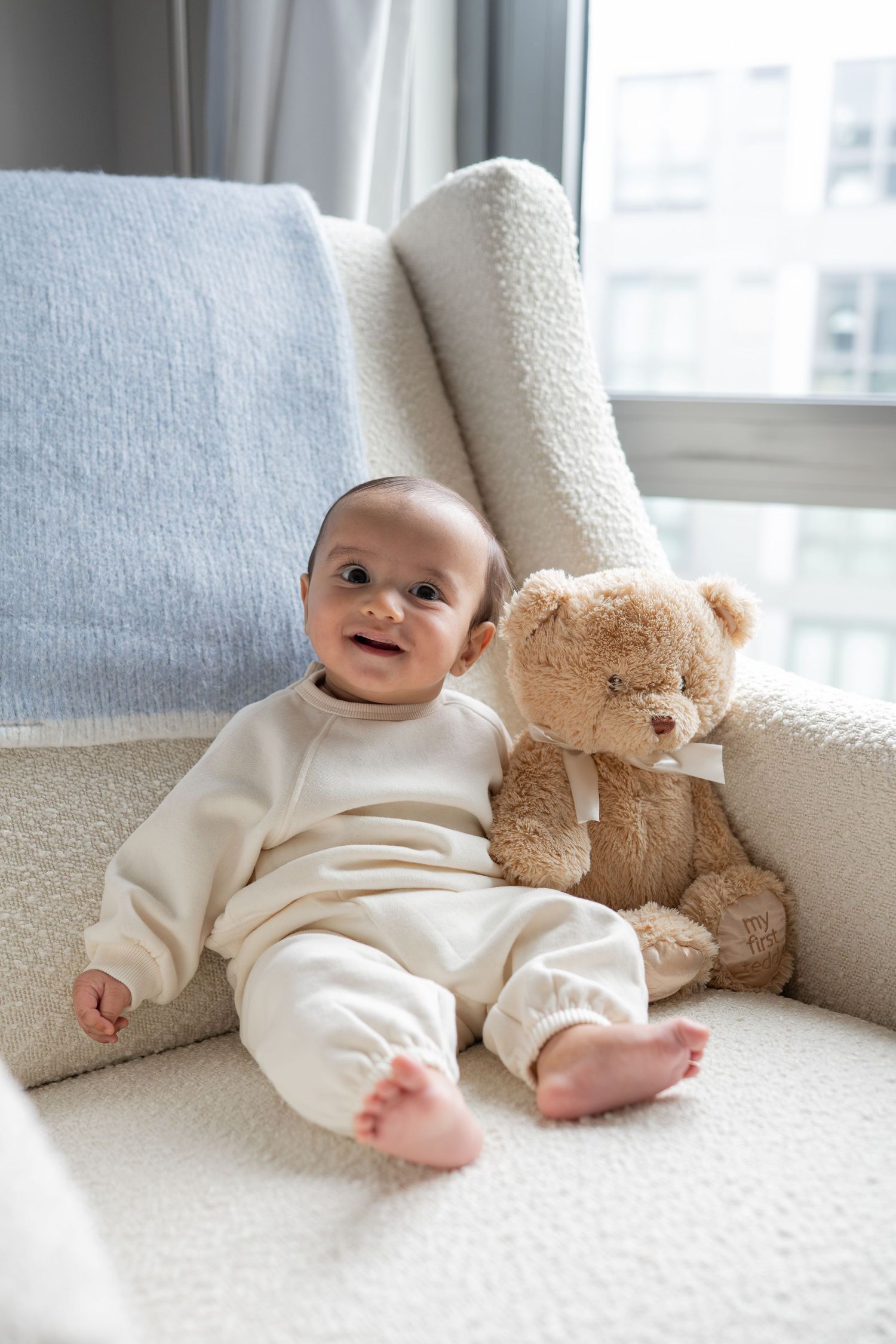A baby in white sleepwear sits on a light-colored couch with a brown teddy bear in a series of sweet moments.