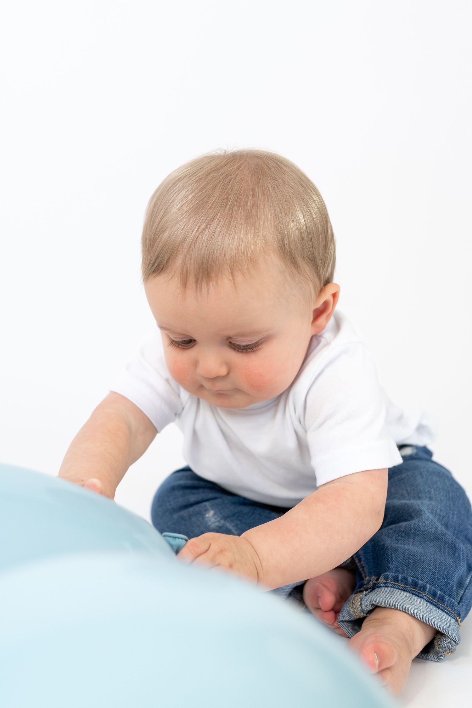 Baby in white tee and jeans sitting on white background playing with a light blue balloon.
