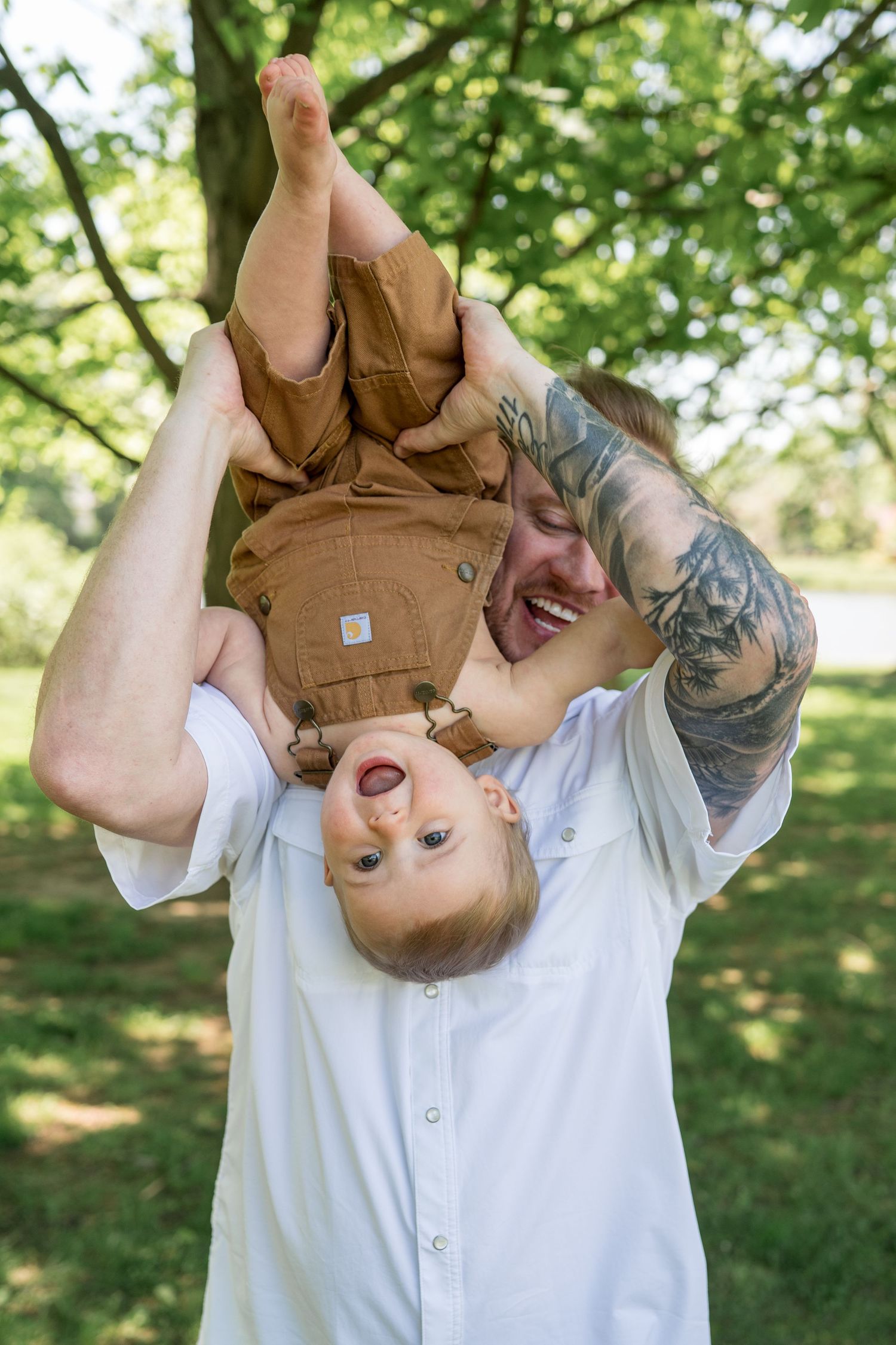 Tattooed father playfully holding baby upside down outdoors, both laughing in a park.