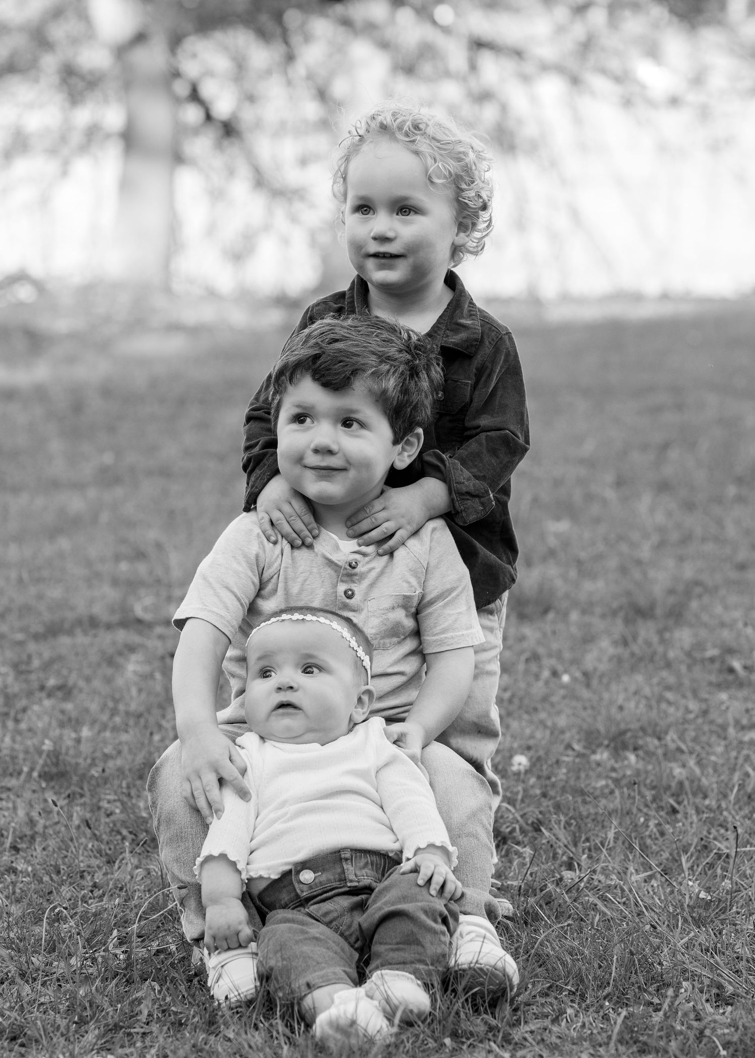 Black and white photo of three young siblings posing together on grass in a park, stacked portrait style.