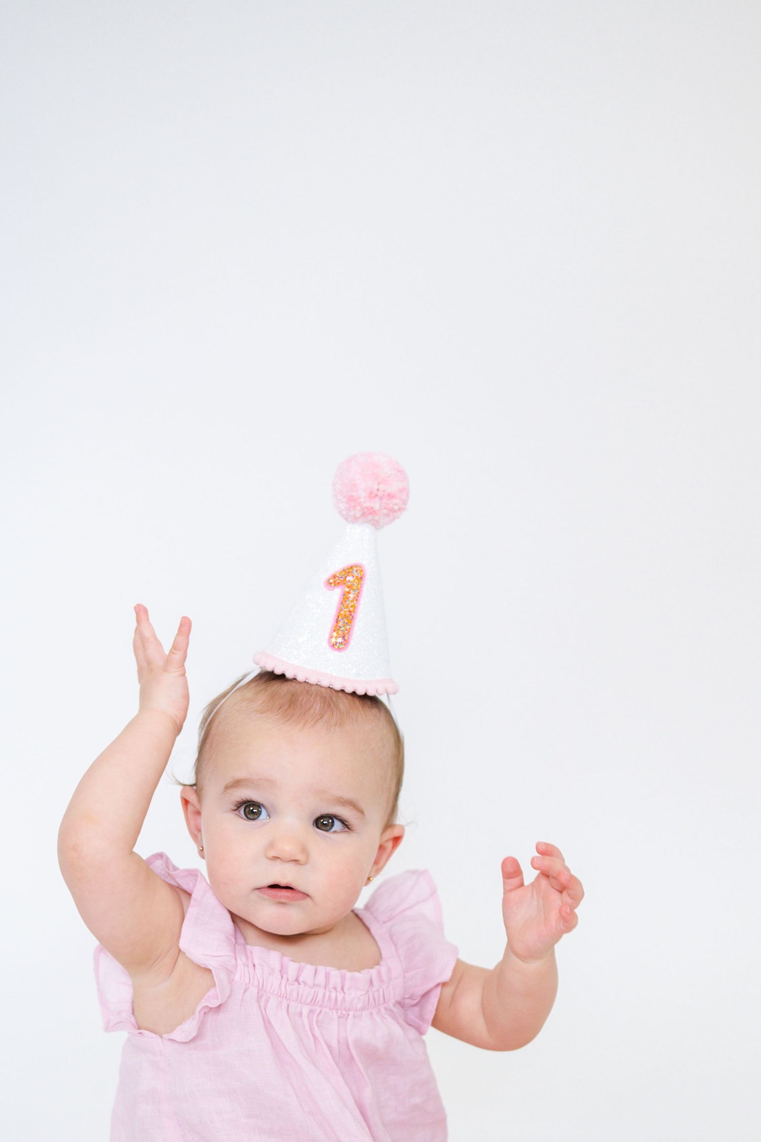 Baby in pink dress raises arms playfully against white background.