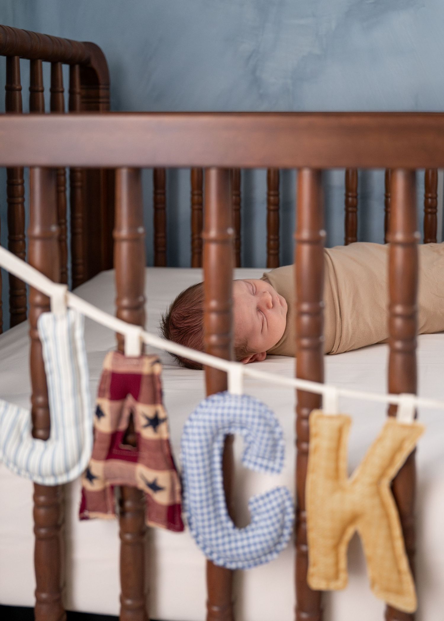 Decorative fabric letters spelling out 'LUCKY' hang from a wooden crib railing against a light background.