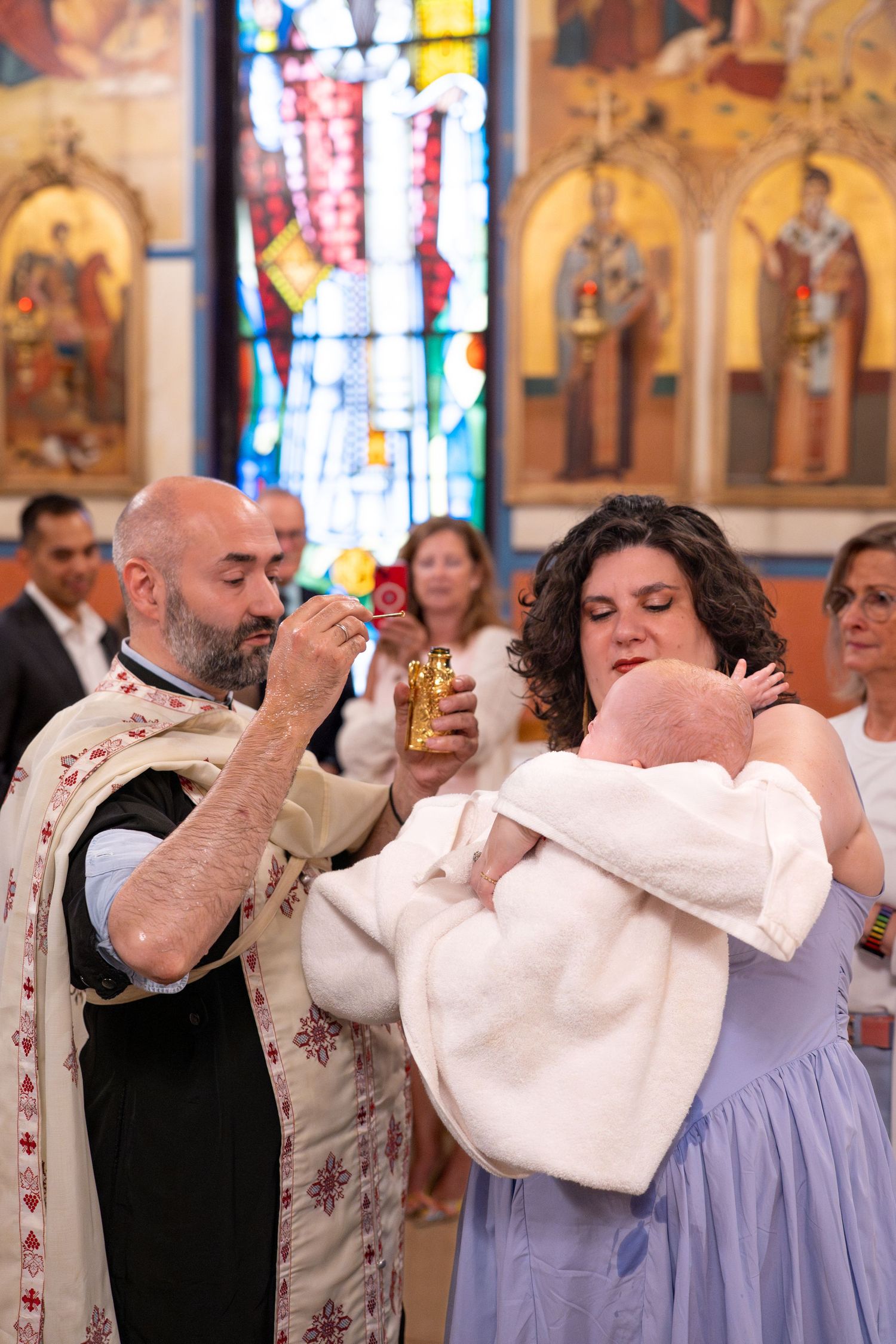 A religious baptism ceremony is performed inside a church with colorful stained glass windows and iconography.