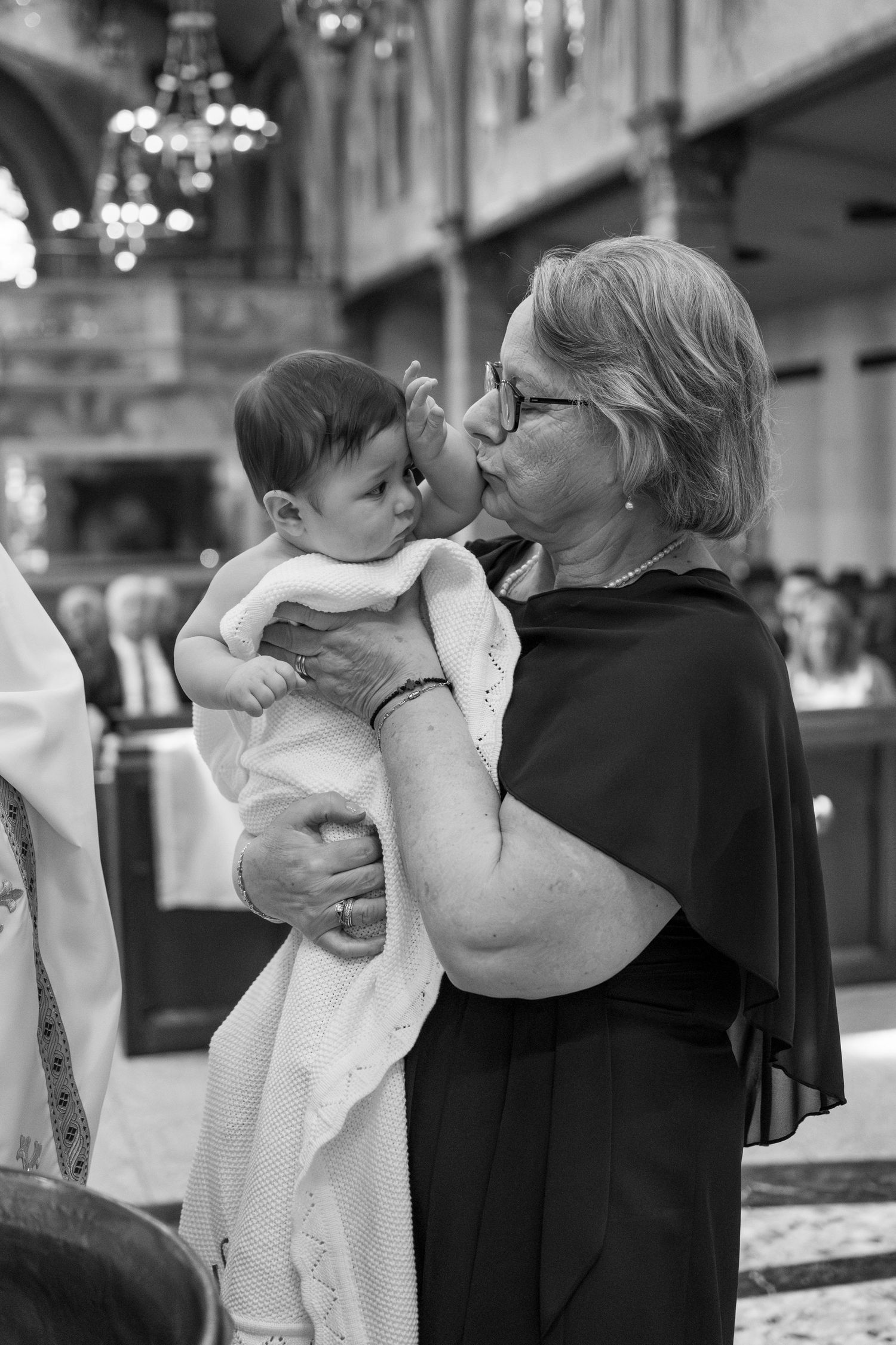 A baby in white baptismal clothing is held during a church ceremony with ornate decorations visible.