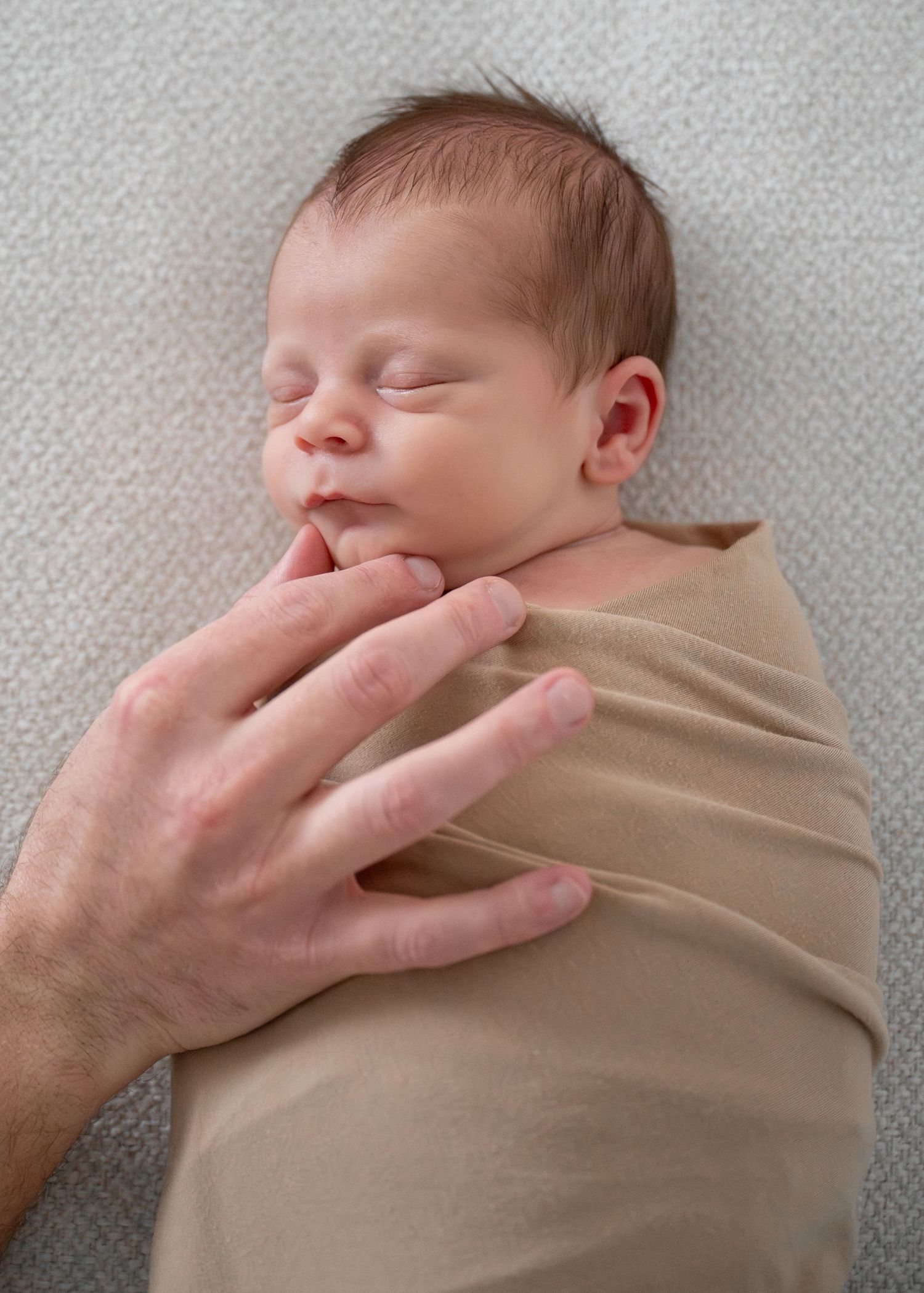 A sleeping newborn baby swaddled in beige fabric with a gentle hand touching their face.