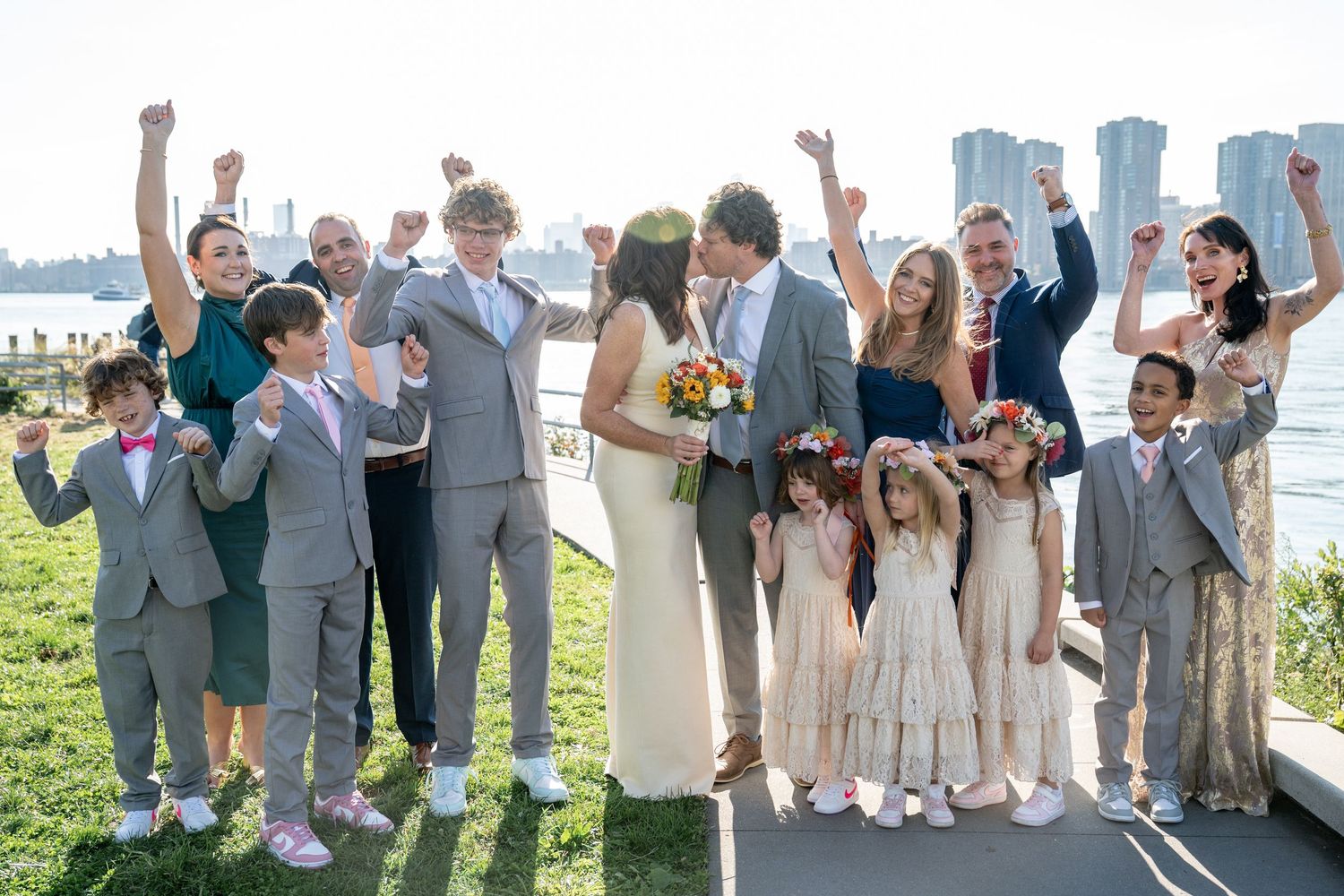 Wedding party members in gray suits and light dresses celebrate joyfully outdoors on a sunny day.