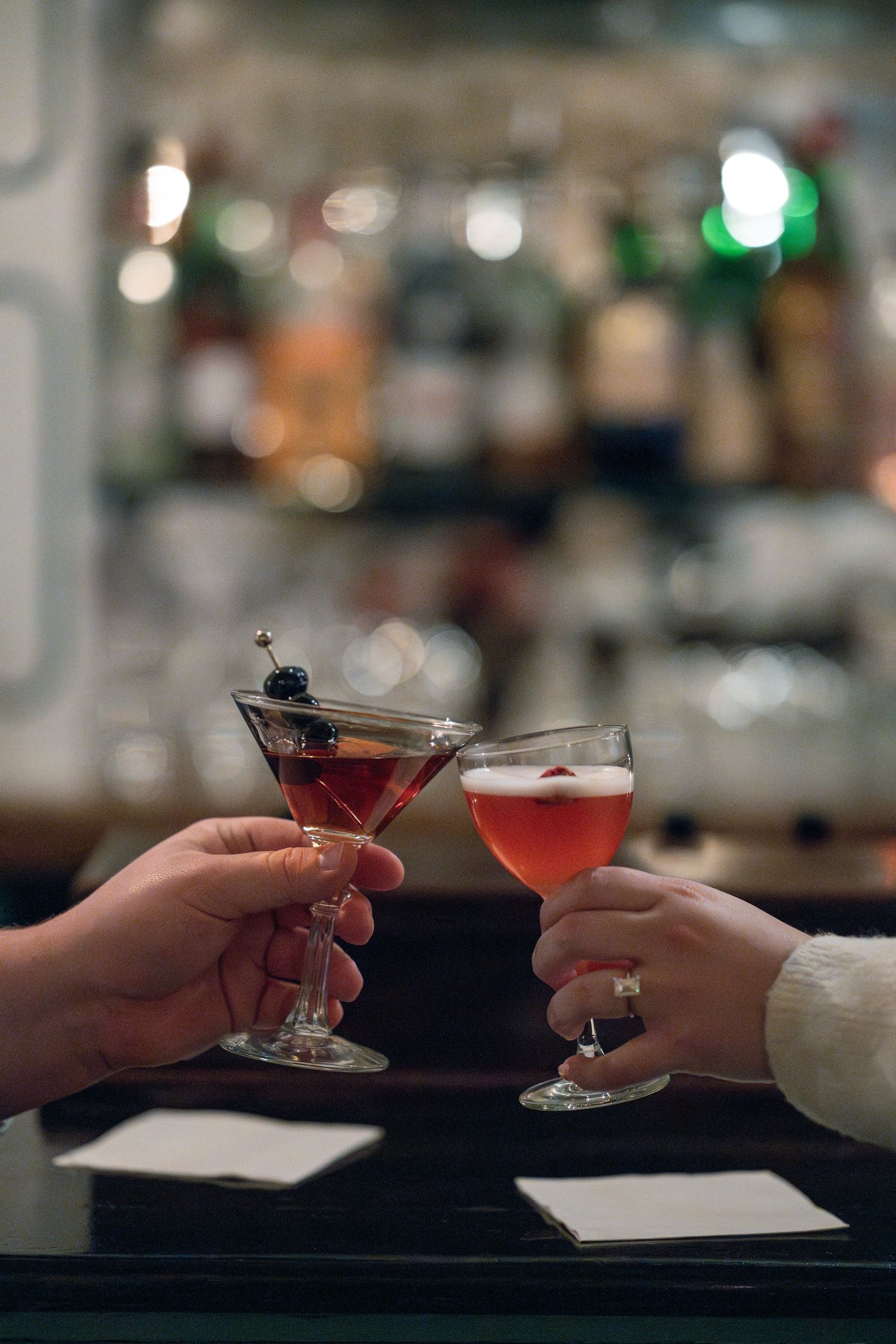 Two hands toast with colorful cocktails at a dimly lit bar celebrating a special moment.