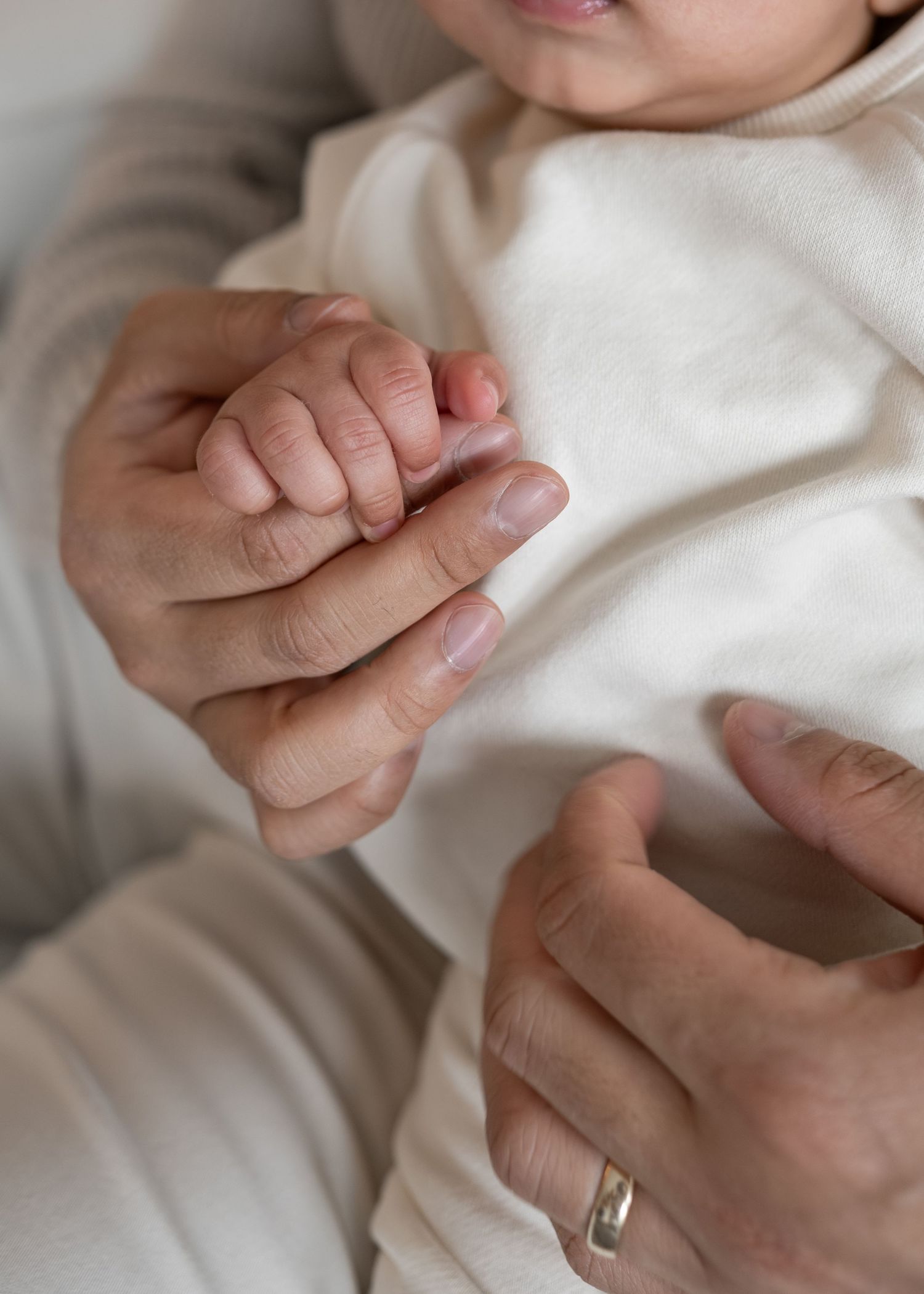 Close-up of newborn baby grasping adult finger with tiny hand, parent wearing gold wedding ring.