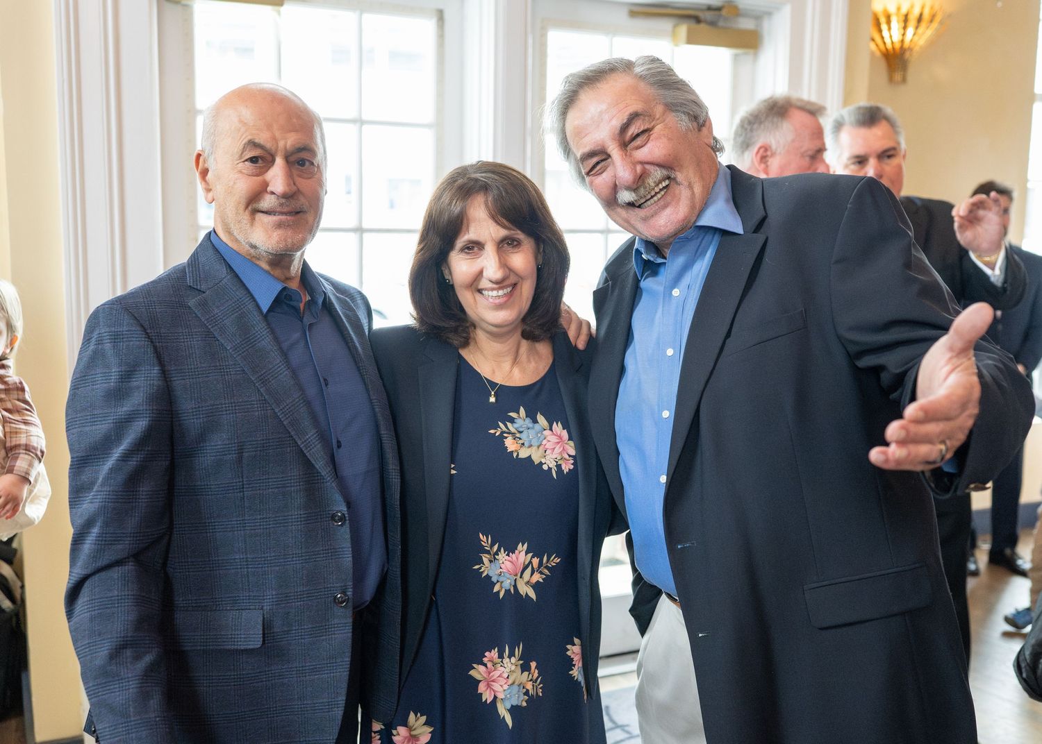 Three smiling adults pose together at a formal indoor gathering, with the man on the right gesturing welcomingly.