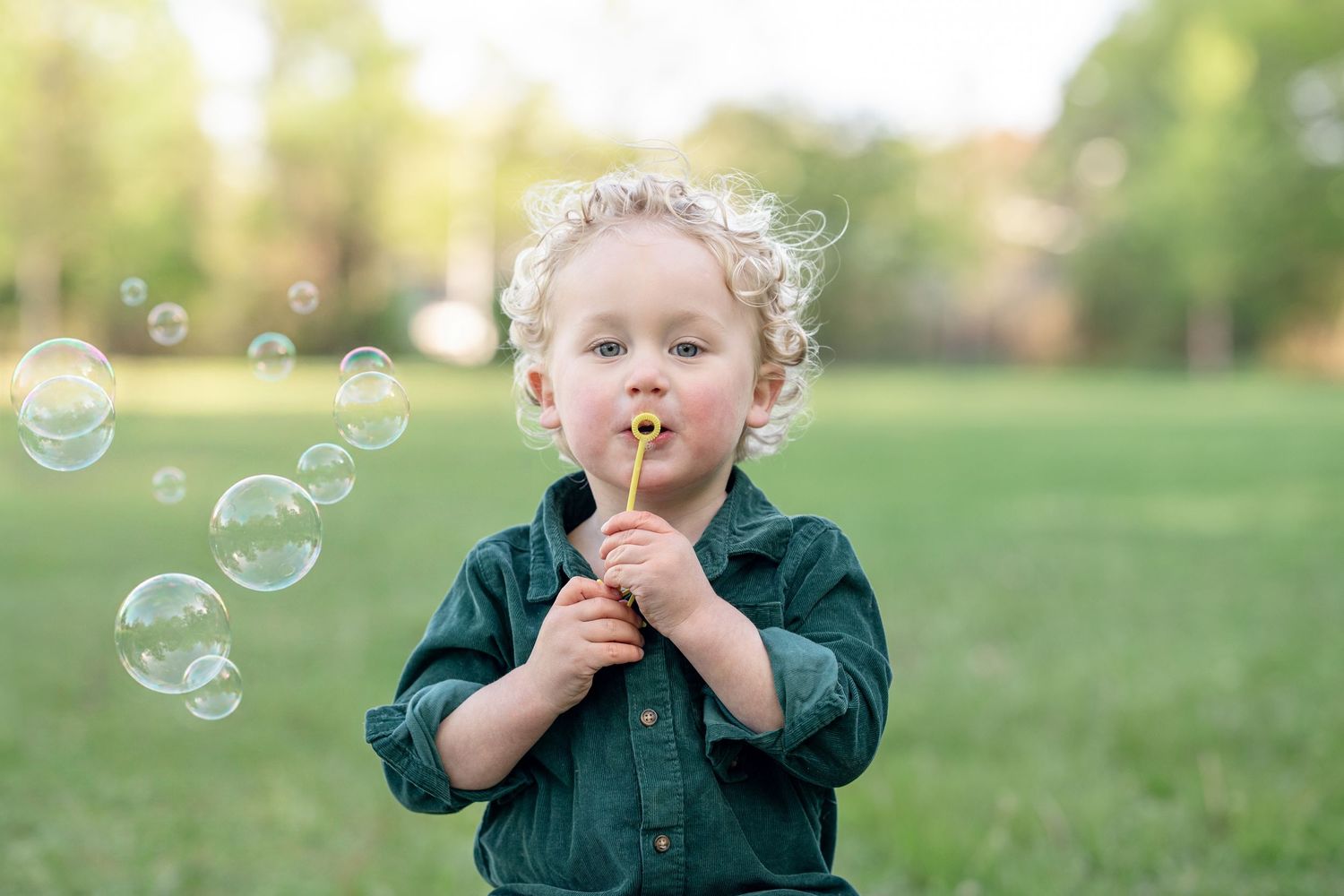 Curly-haired toddler in green shirt blowing bubbles outdoors in a sunny park.