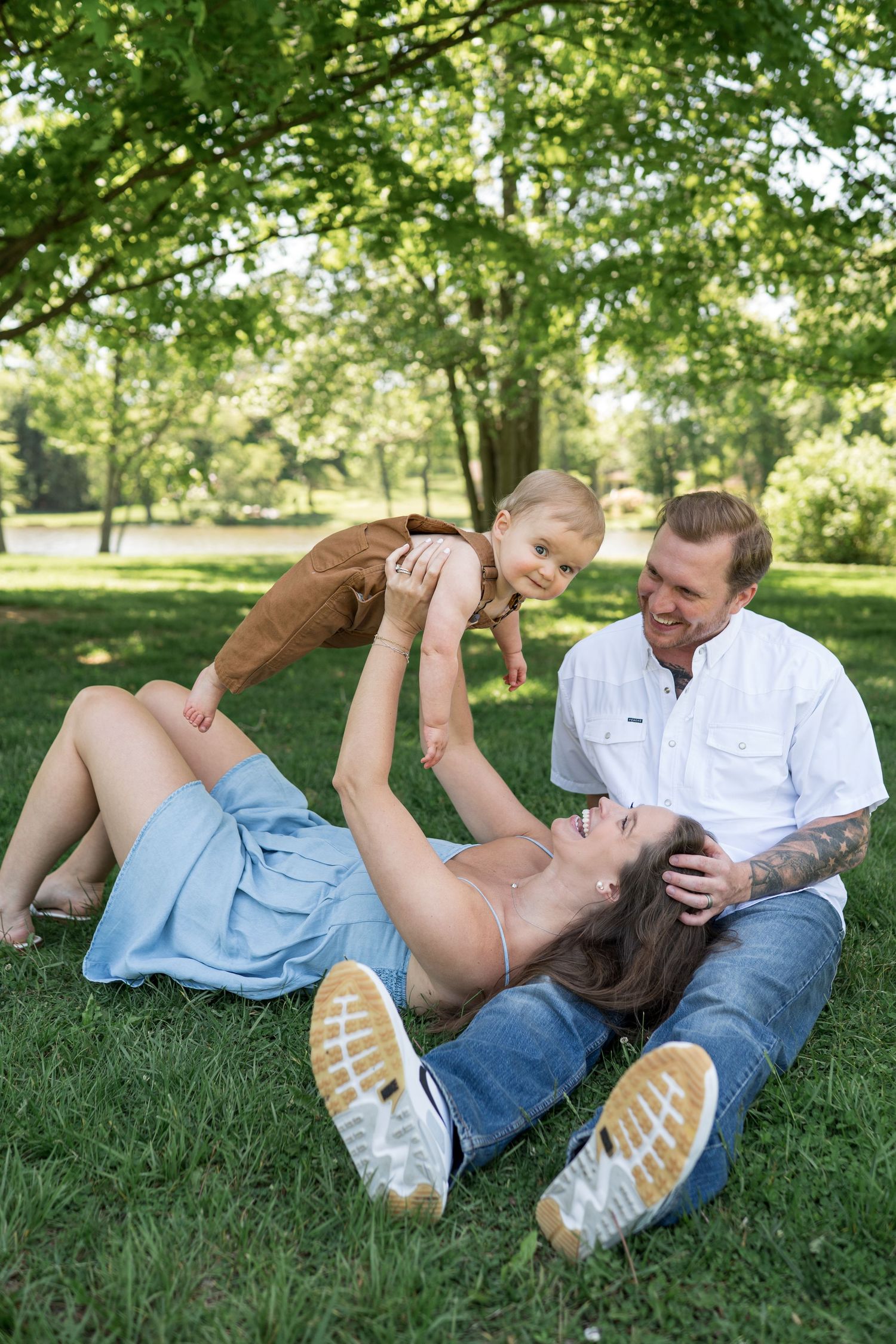 Happy family relaxing in green park, mother lifting toddler while tattooed father smiles on sunny day.
