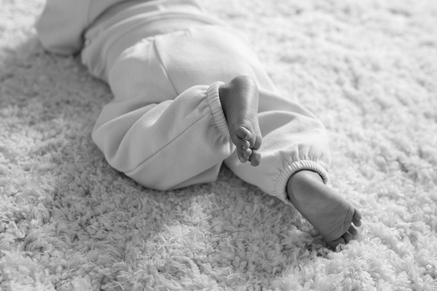 Black and white photo of a baby lying on a fluffy white carpet, wearing white pants with bare feet visible.