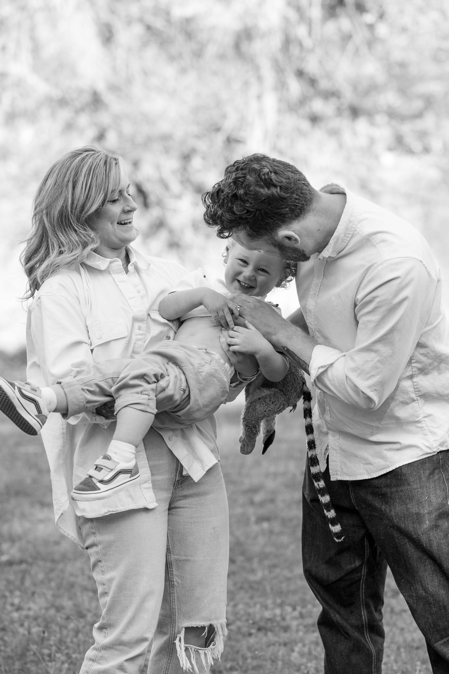Black and white photo of happy parents laughing with their joyful baby outdoors in a park setting.