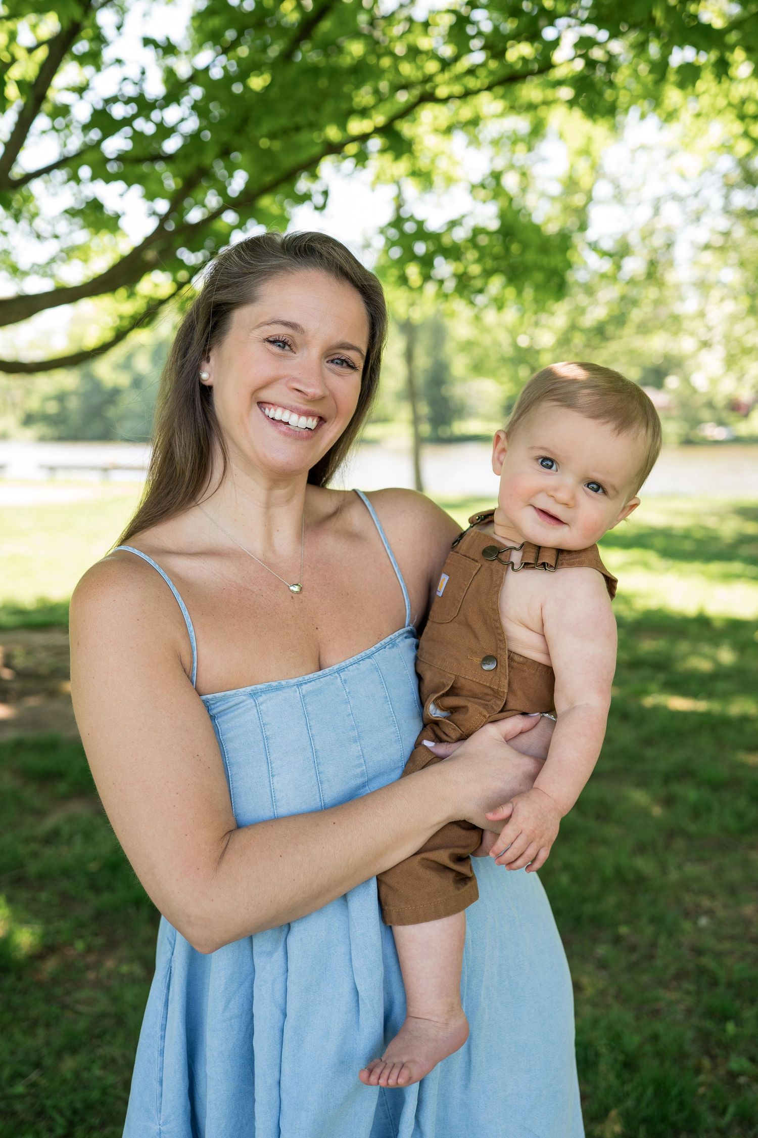 Smiling woman in blue dress holding a baby in brown overalls outdoors in a green park.