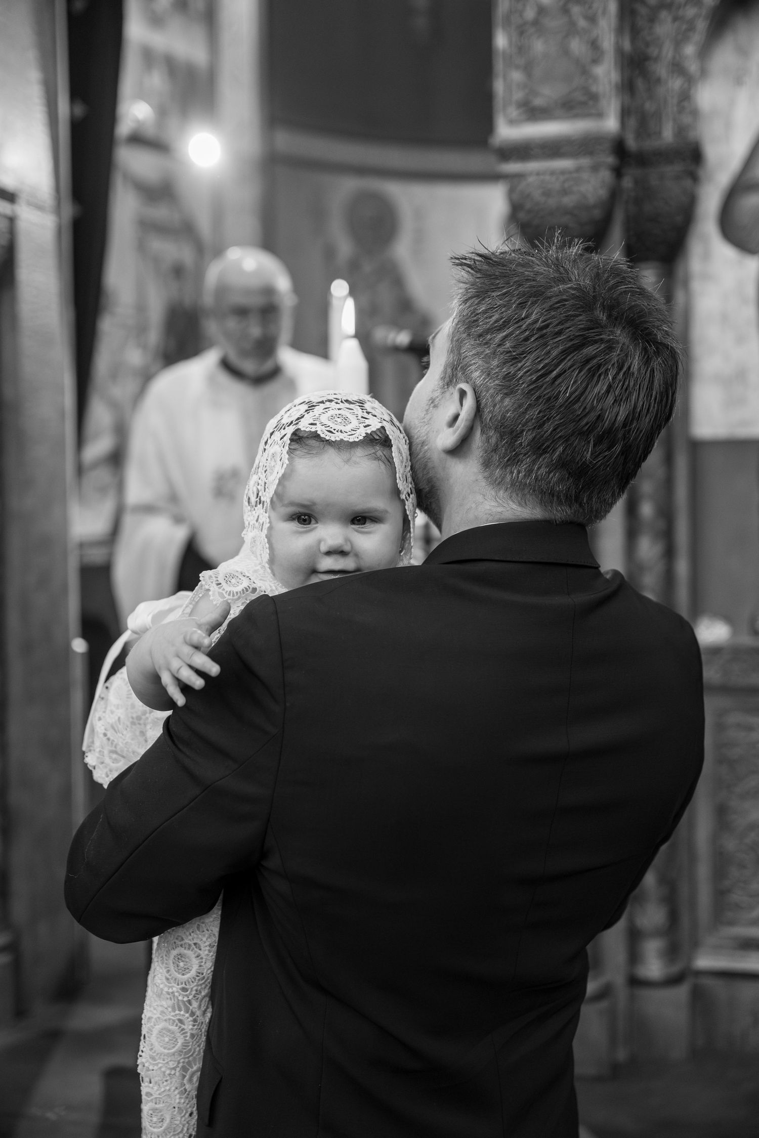 Black and white photo of a man holding a baby in a christening gown inside a church with a priest in the background.