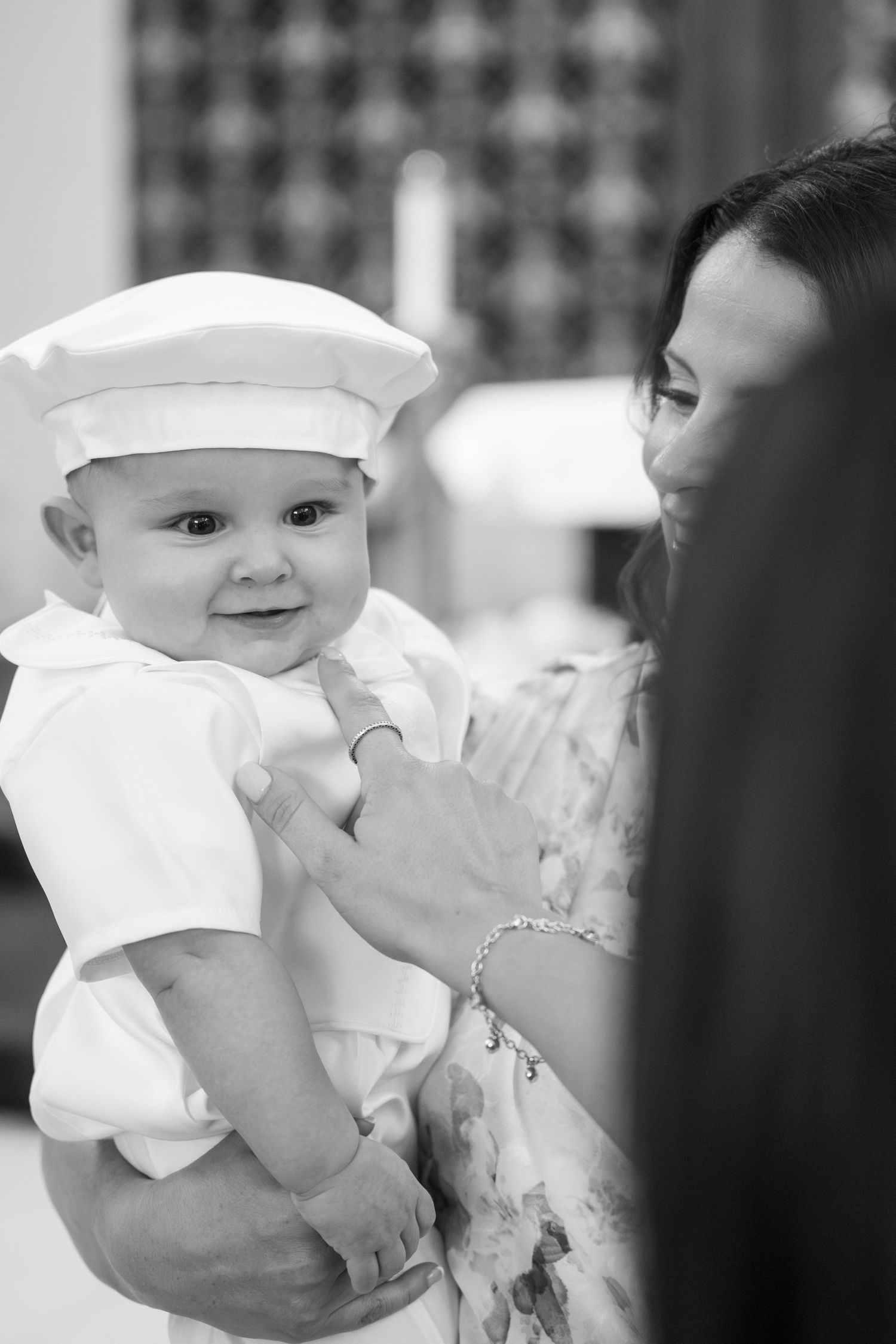 A baby in a white christening outfit and cap being held at a formal event.
