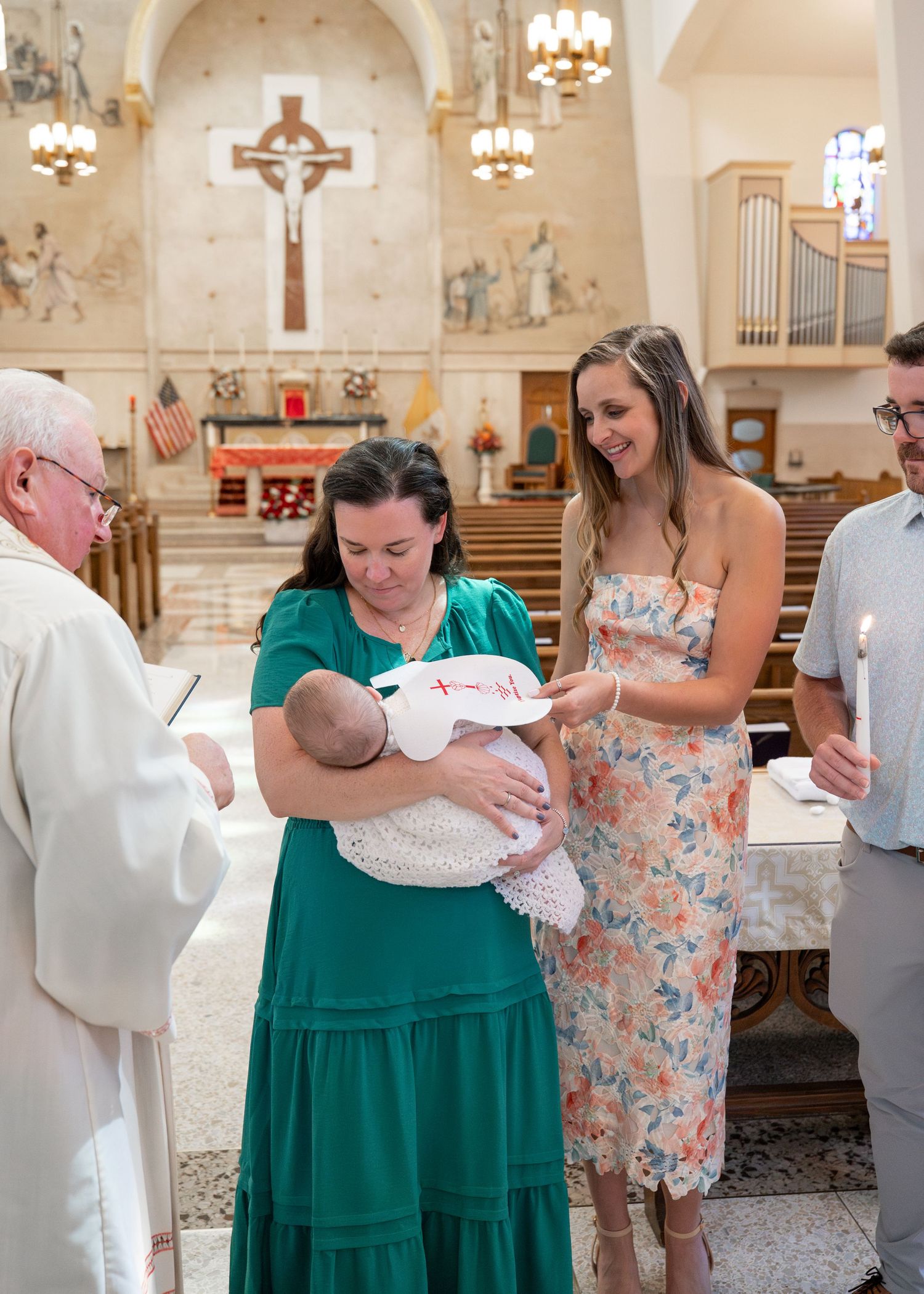 A baptism ceremony taking place inside a Catholic church with a large crucifix and stained glass windows visible in the background.