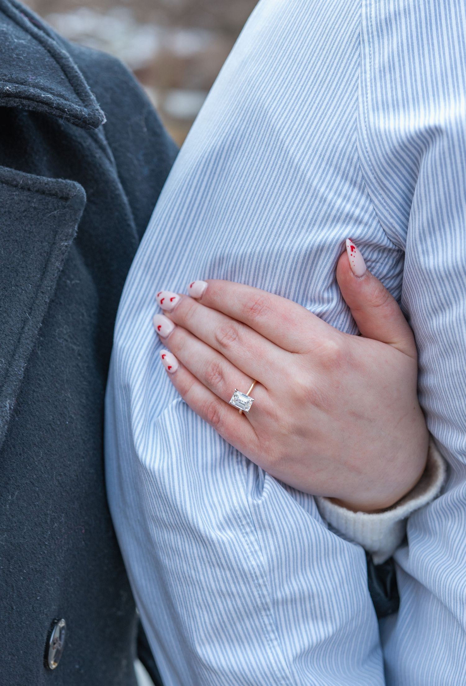 Close-up of an engagement ring being shown on a hand against a light blue striped shirt.
