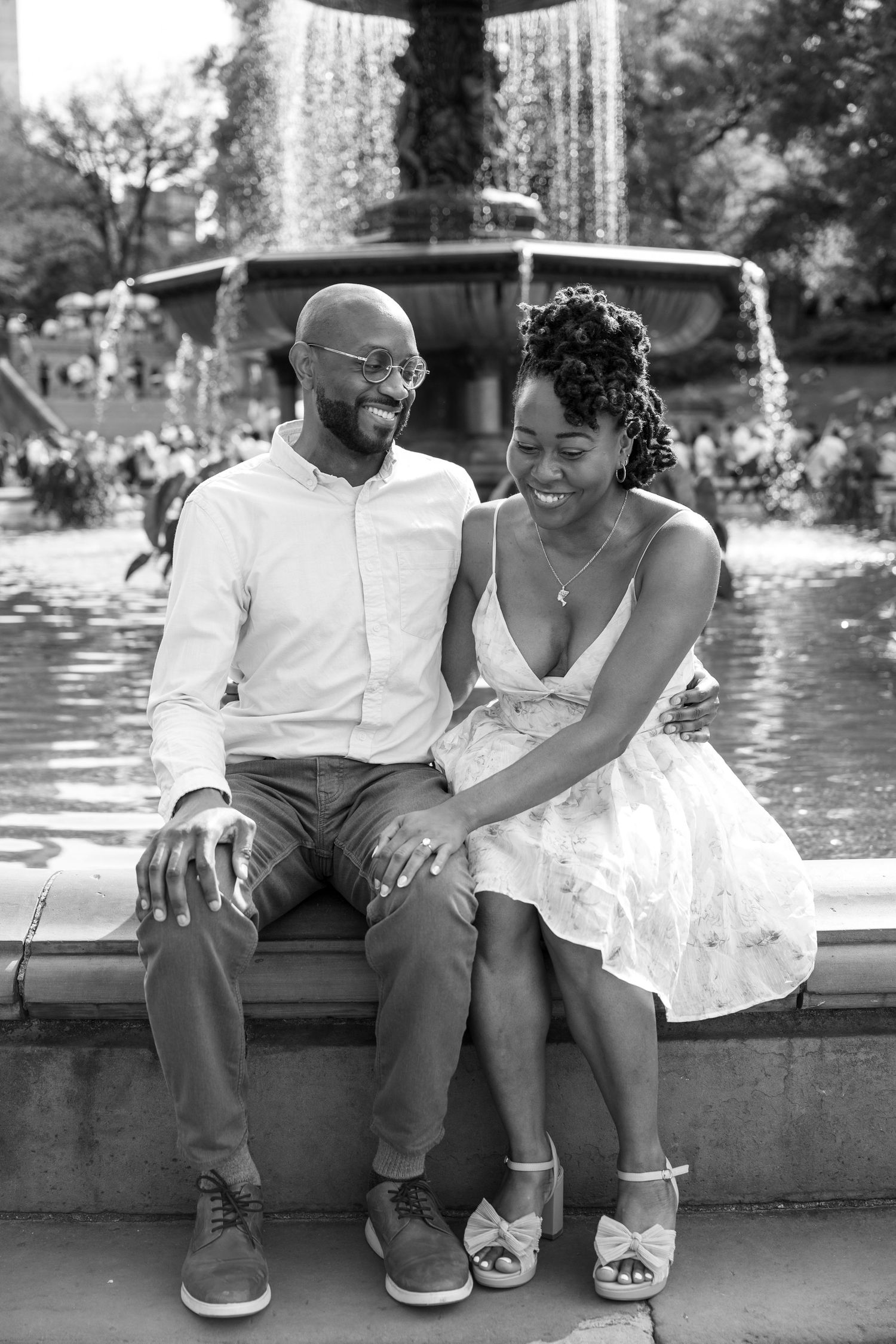 A couple shares a joyful moment together while sitting on a fountain ledge in a park on a sunny day.